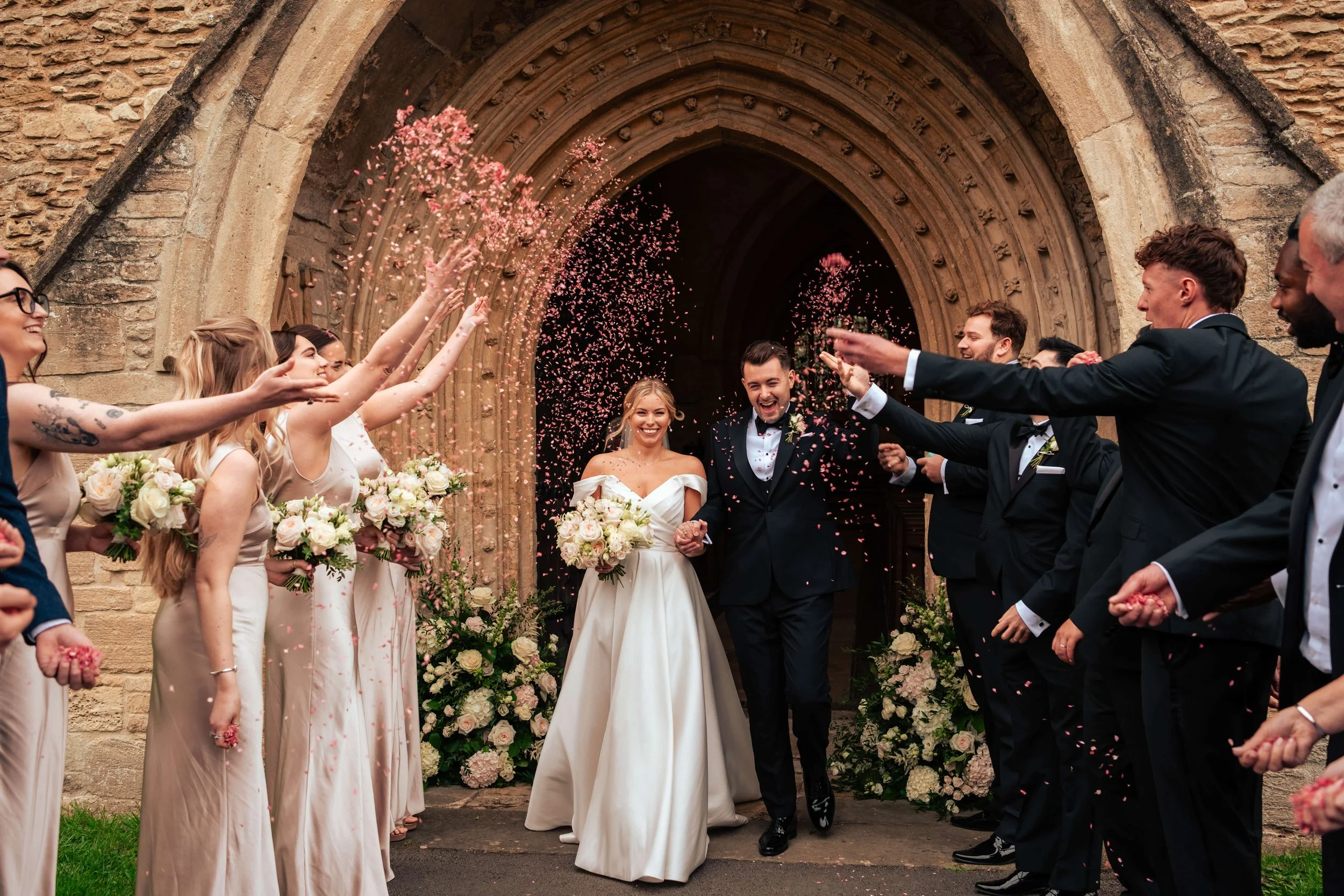 A newlywed couple walking out of a church surrounded by friends and family throwing pink confetti, with floral arrangements on either side of the church entrance.