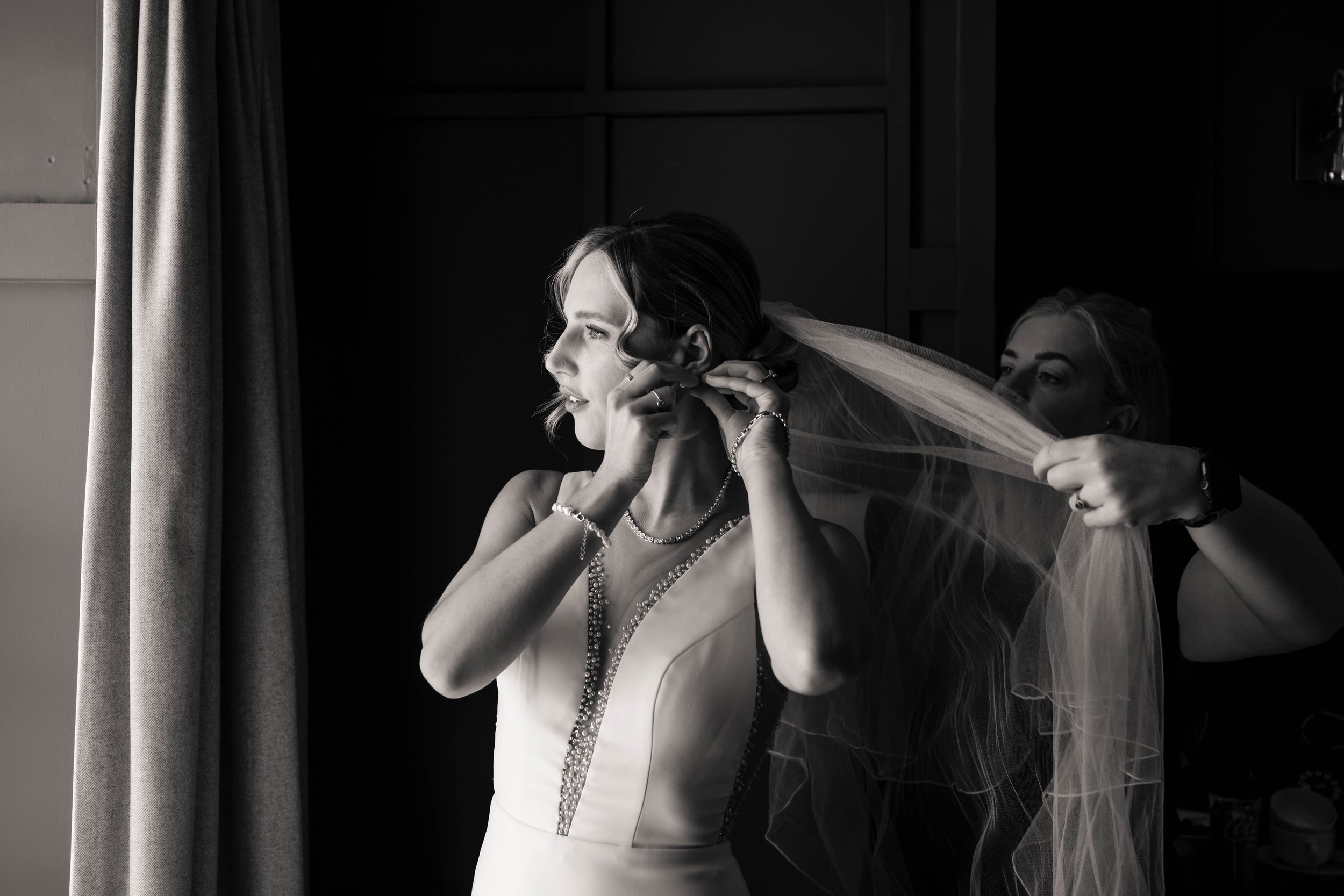 Bride putting on her earrings in the morning of her wedding beside a well-lit window and curtain.