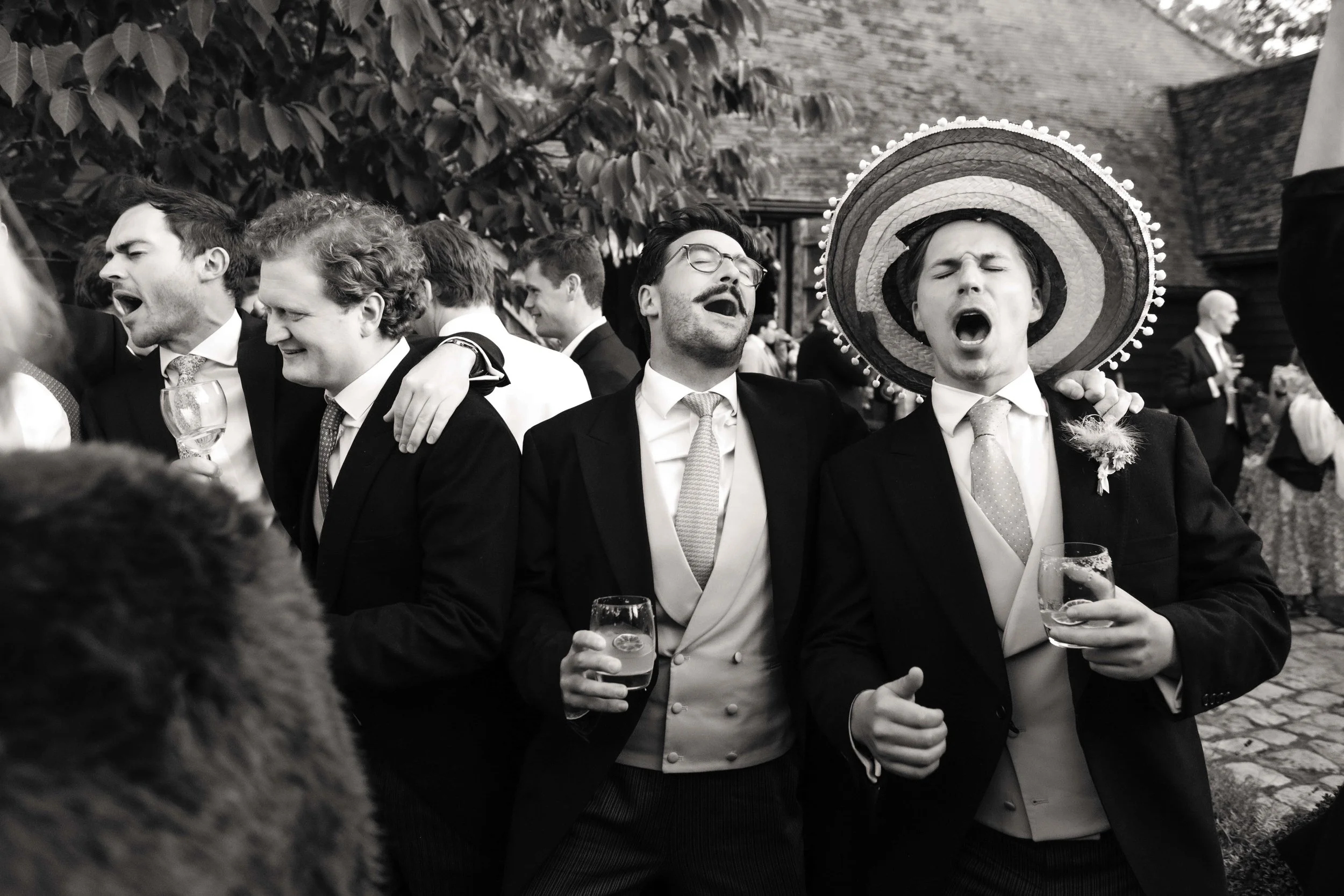Group of men in suits celebrating at a wedding, singing and drinking wine, one wearing a sombrero.