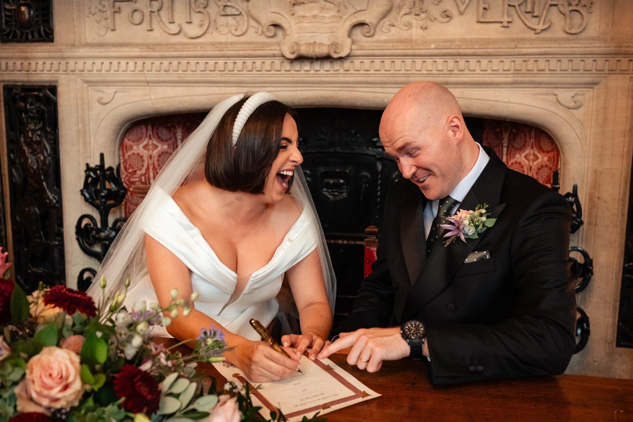 A bride and groom are signing a marriage license at a wedding ceremony, laughing and sharing a joyful moment in front of a fireplace with decorative woodwork.