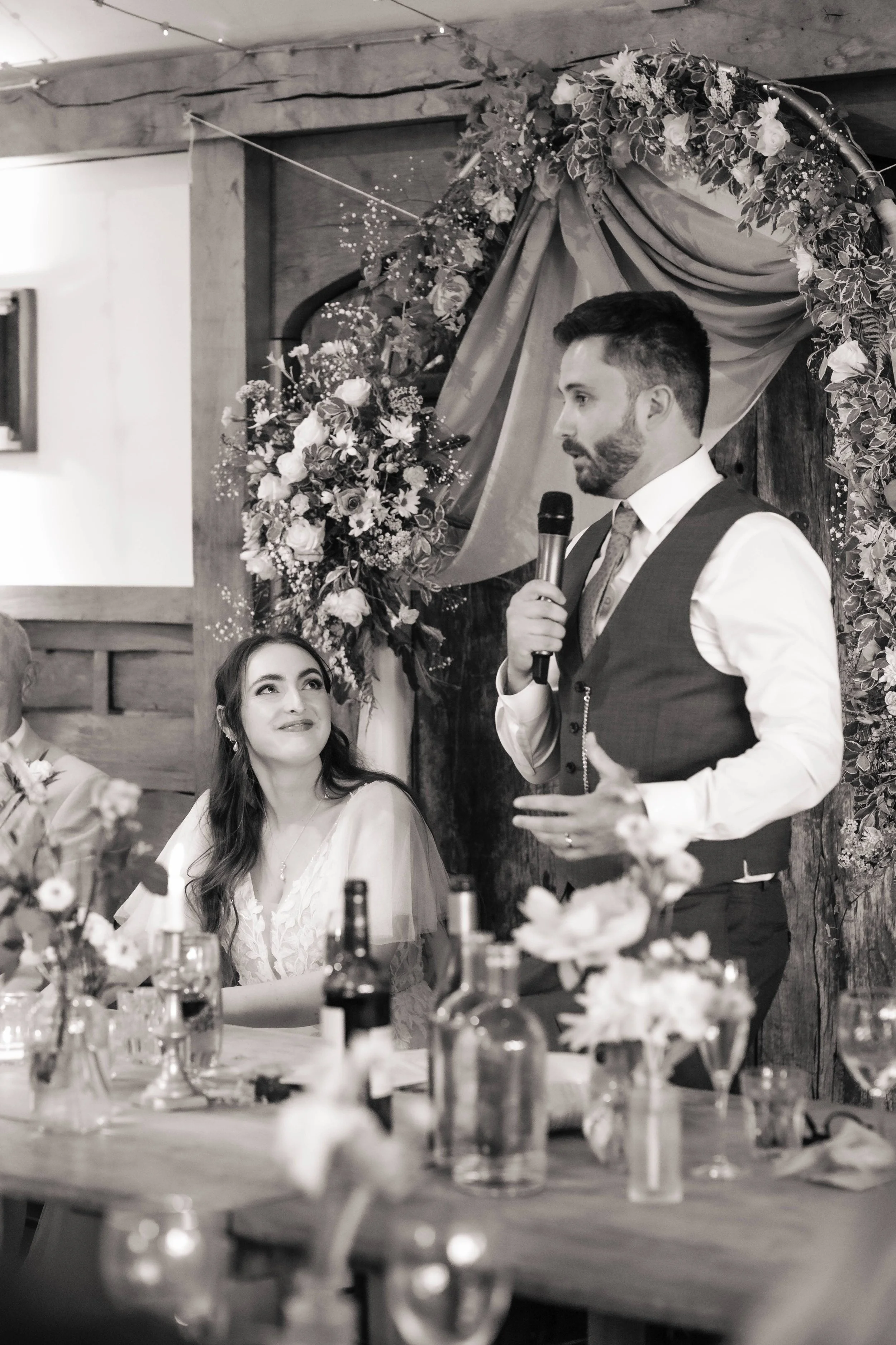 A man giving a speech at a wedding reception, holding a microphone and standing next to a seated woman who is looking up at him. The background features floral decorations and draped fabric.