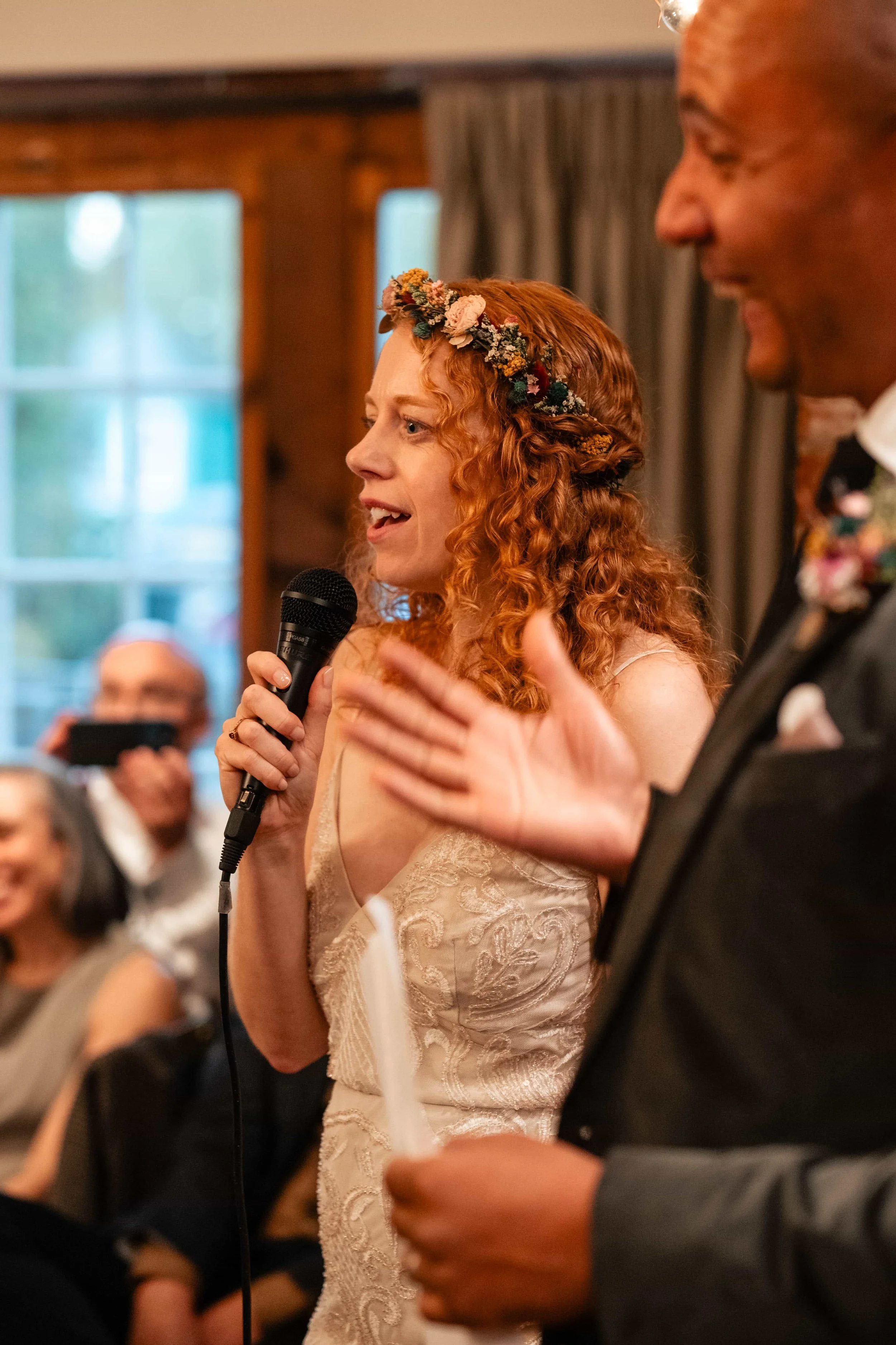 A red-haired woman with a floral headband is speaking into a microphone during a wedding reception, with guests seated in the background.