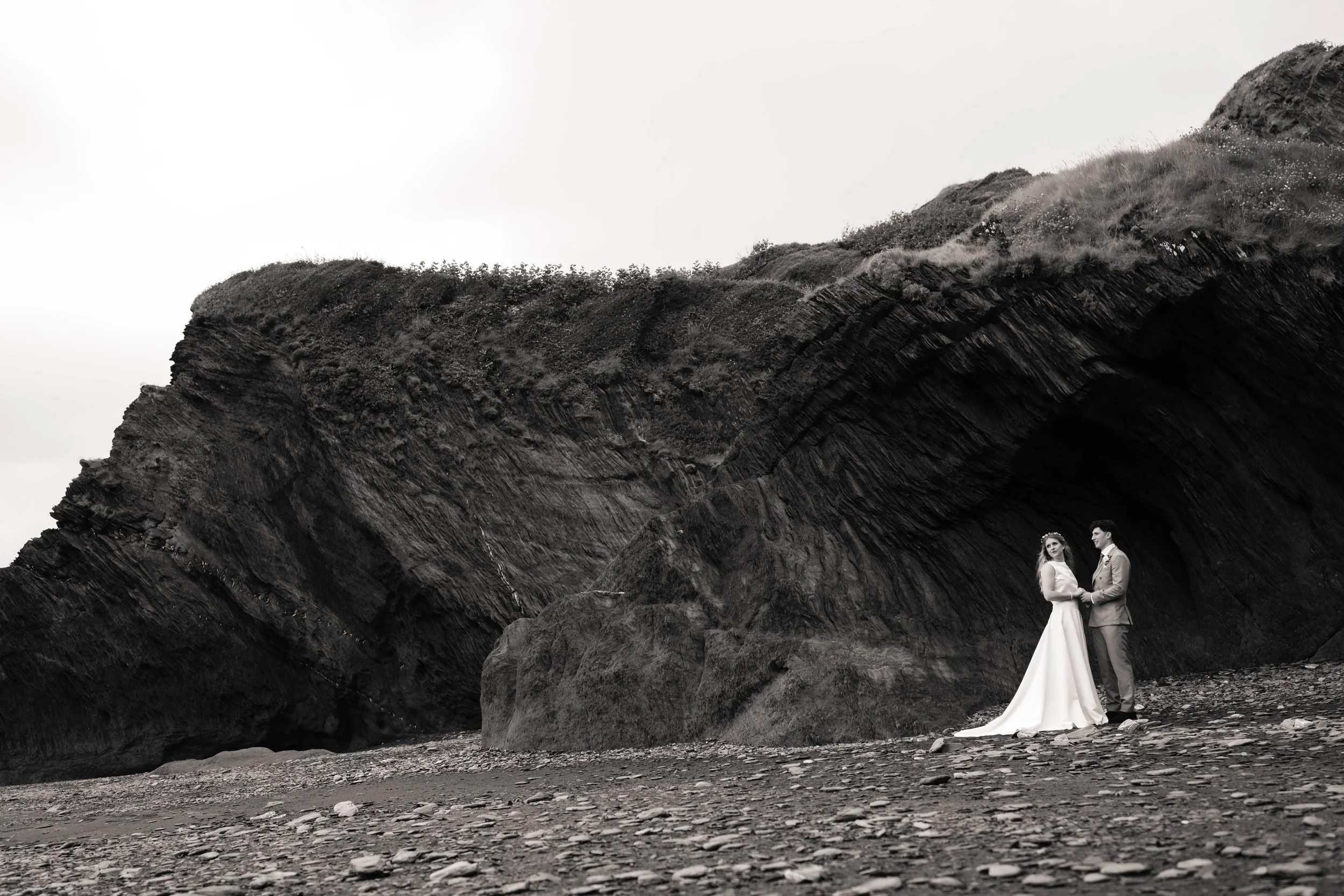 Bride and groom holding hands beside a large cliff, with the bride looking out along the beach.