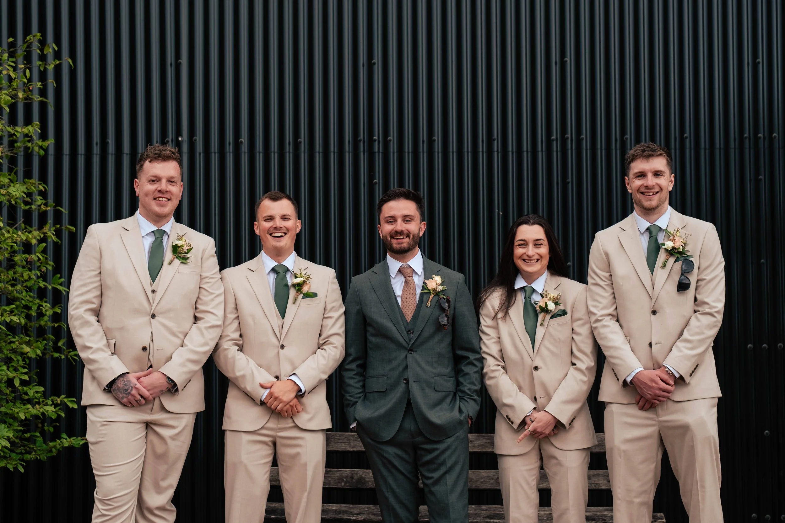 Groom and groomspeople in formal suits standing outdoors against a black corigated wall, smiling for a group portrait.