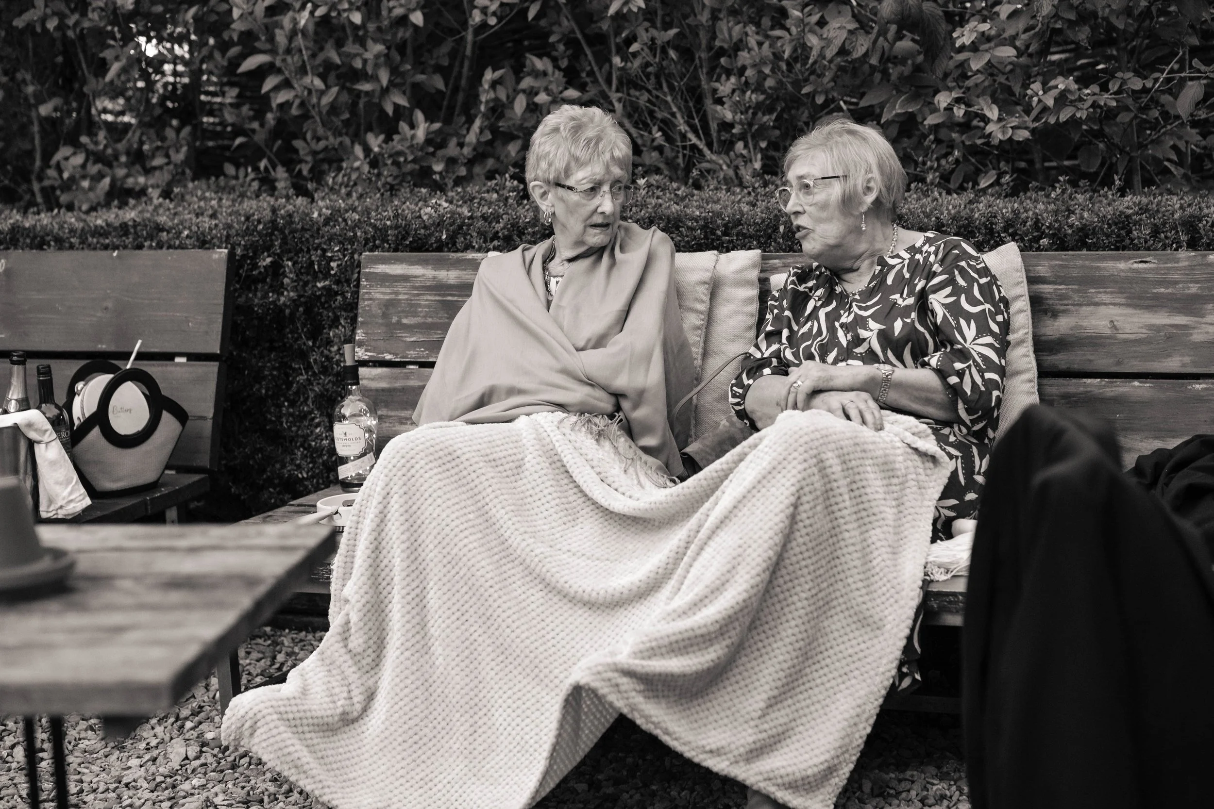 Two elderly women sitting on a wooden bench outdoors, engaged in conversation, with bottles and a purse on a bench nearby, covered with blankets.