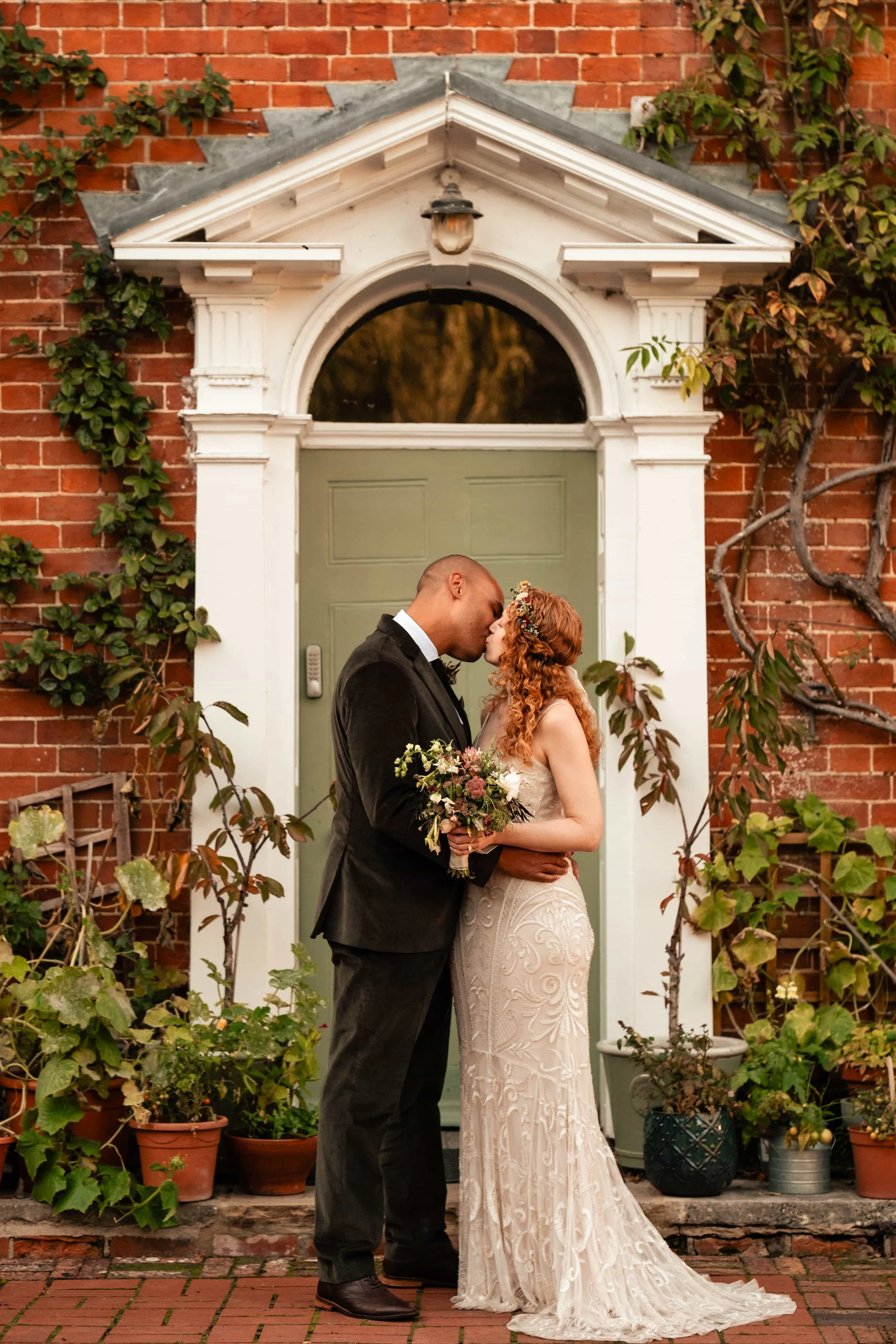 A couple dressed in wedding attire sharing a kiss in front of a house door. The woman has curly red hair and is holding a bouquet of flowers, while the man is wearing a black suit. The house has an arched door with white trim and a brick exterior, de