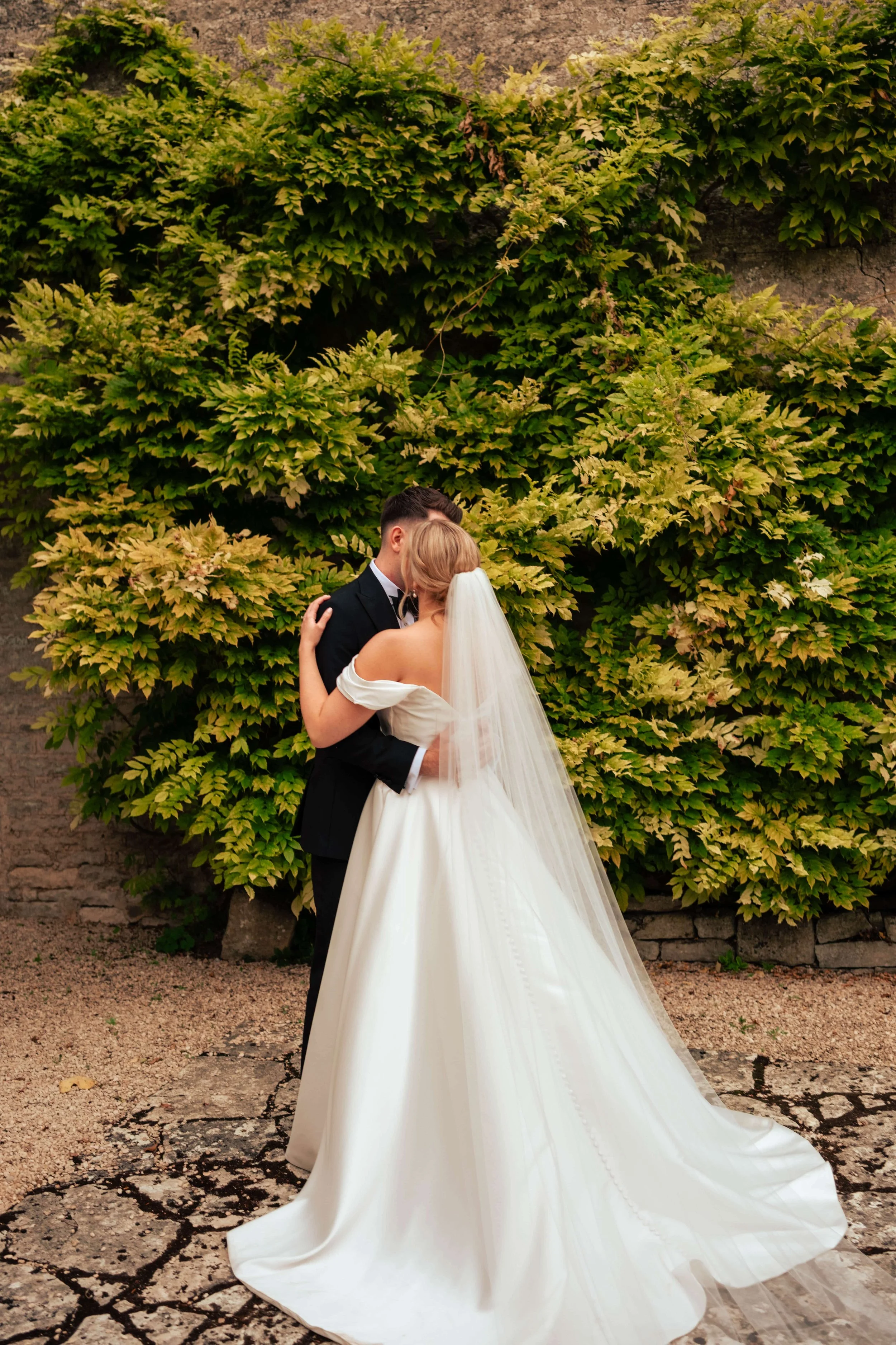 A bride and groom embrace each other outdoors, with a lush green ivy-covered wall in the background.