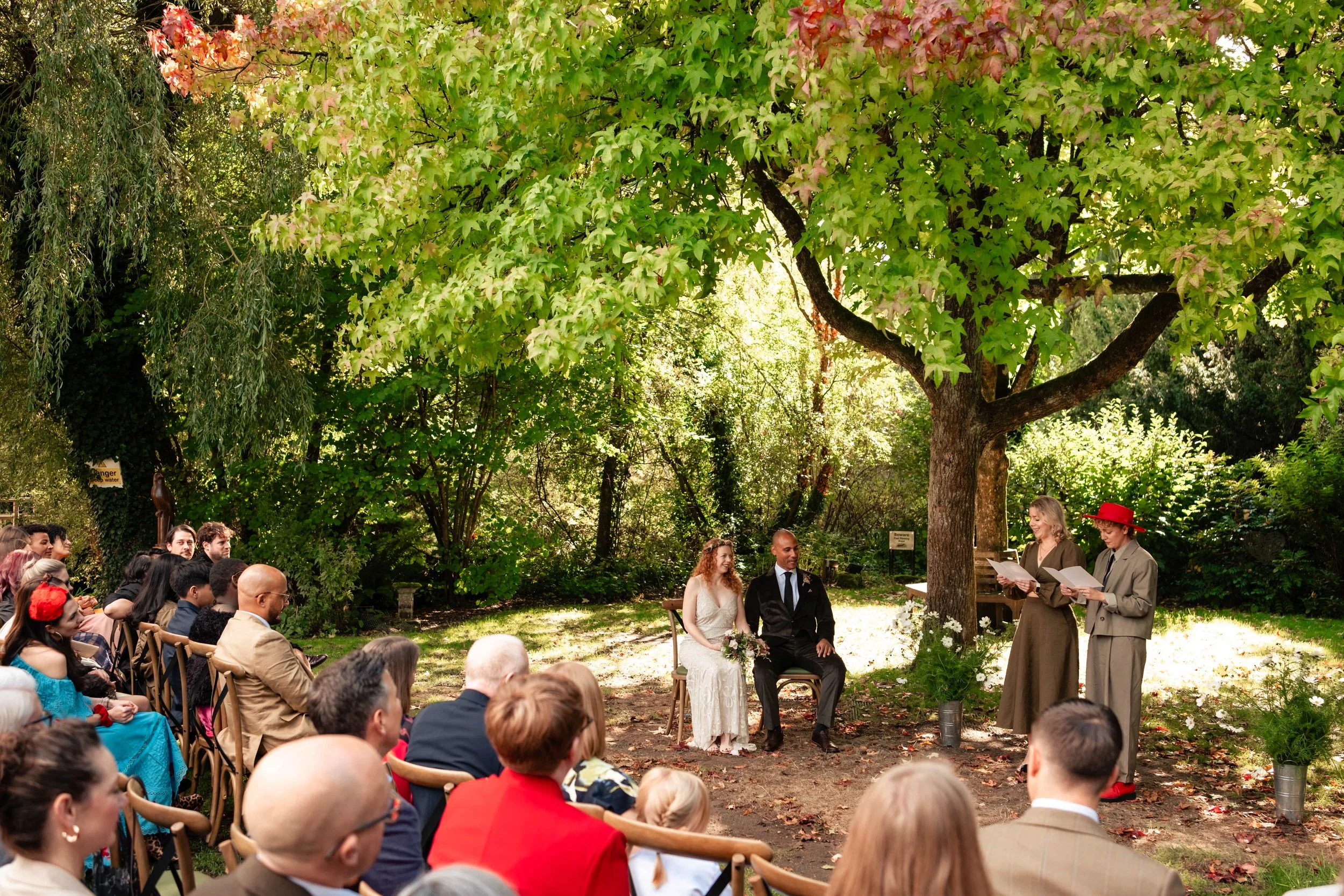 A wedding ceremony outdoors under a large tree with green leaves and sunlight filtering through, featuring a bride and groom sitting next to each other, surrounded by seated guests, with officiants reading from papers.