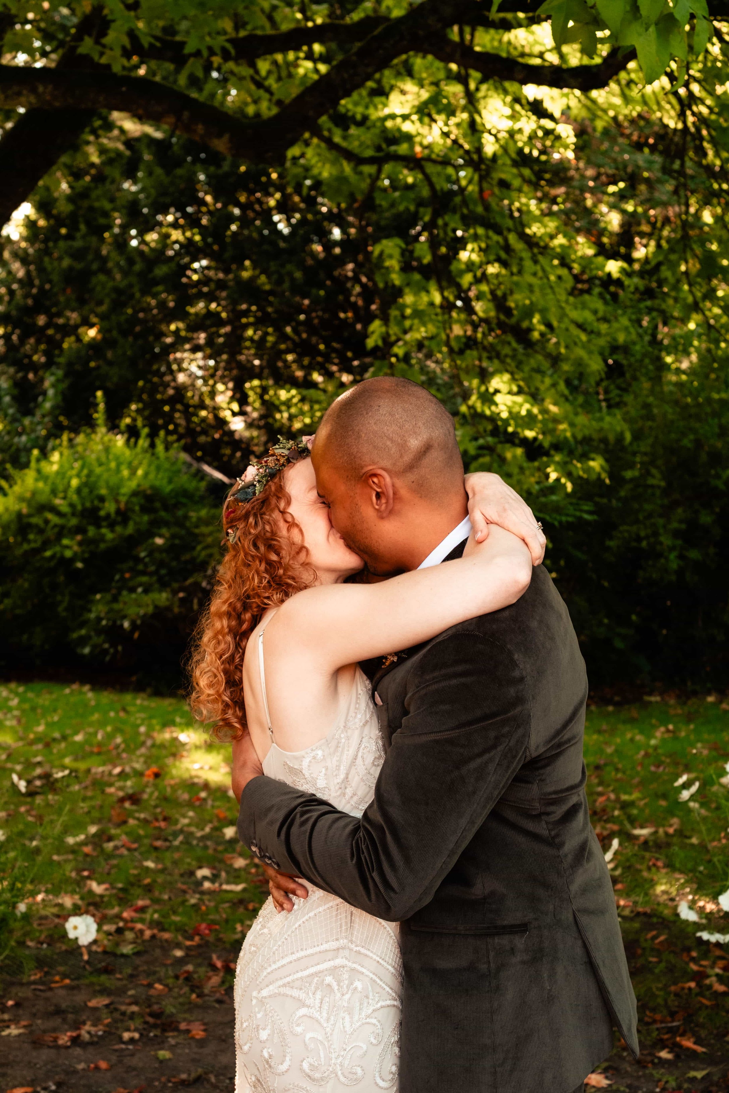 A couple is embracing and kissing outdoors in a lush green park, with trees and fallen leaves in the background.