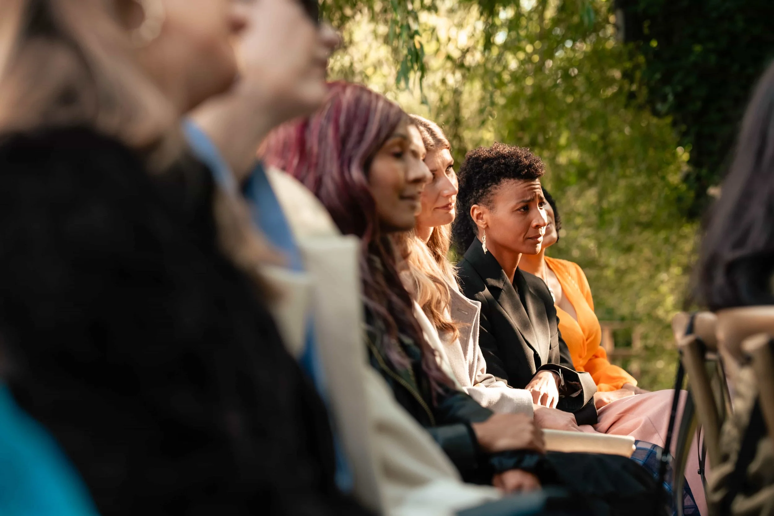 A group of women sitting outdoors, attentively listening during daytime. The setting appears to be a park or garden with green trees in the background.