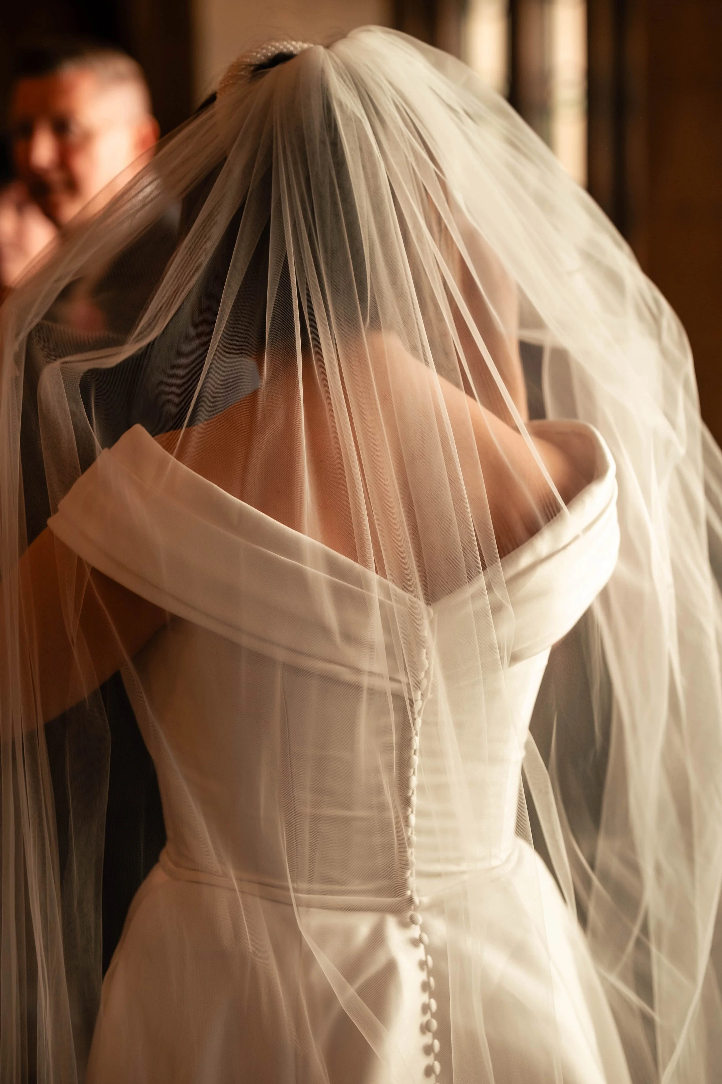 A bride in an off-the-shoulder white wedding dress with a long veil covering her head and face, standing indoors.