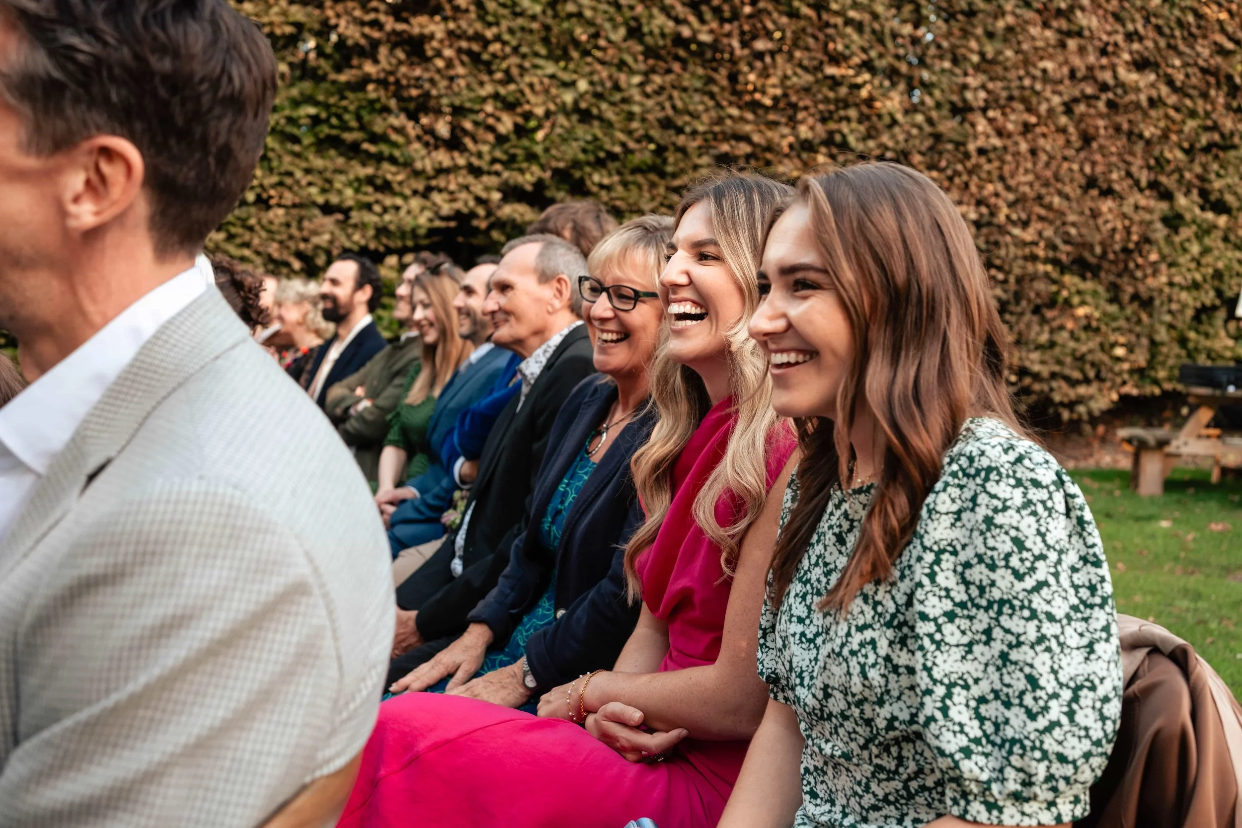 People sitting outdoors, smiling and laughing at an event, with trees in the background.
