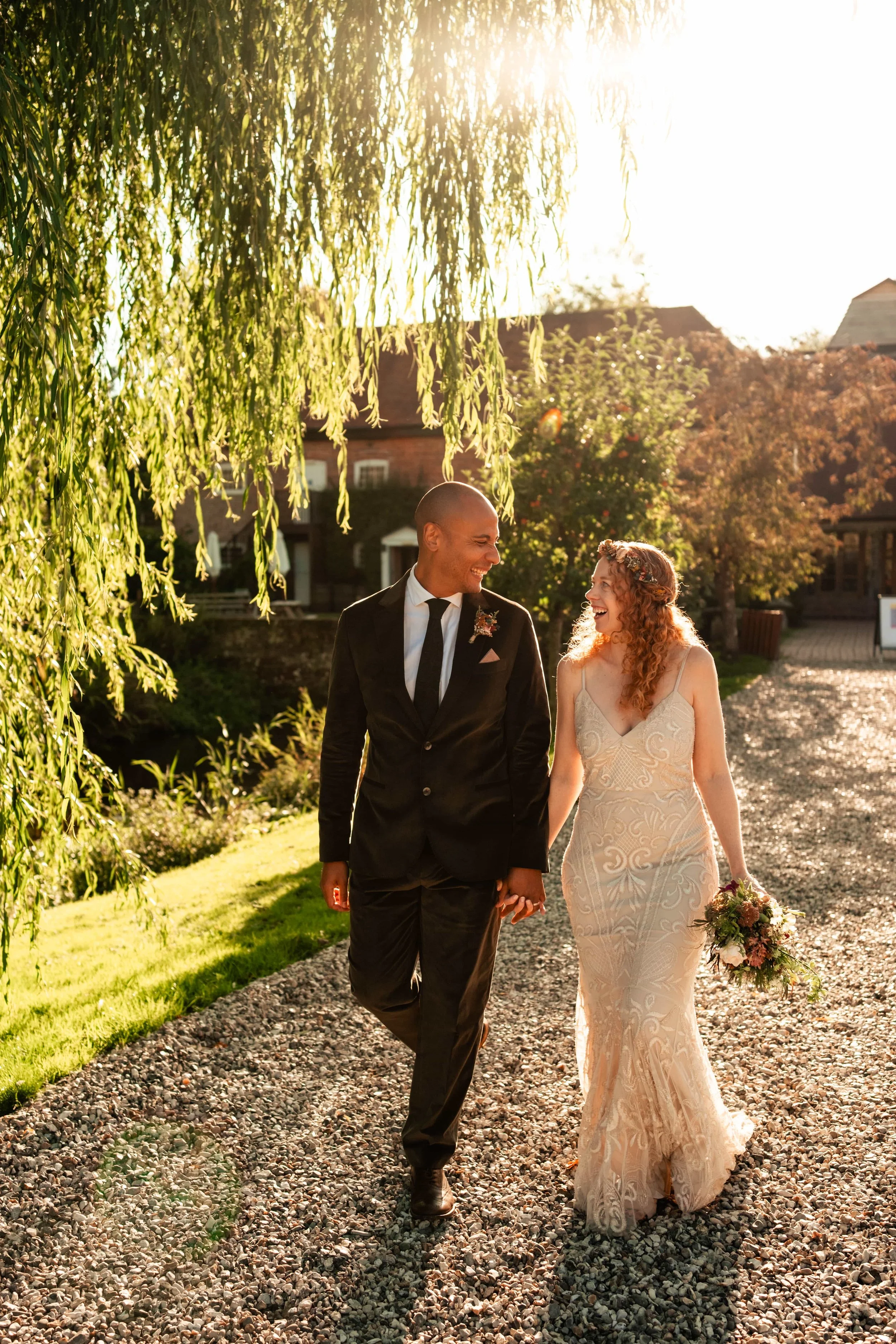 Bride and groom holding hands and smiling, walking outdoors during sunset, surrounded by greenery and a few houses in the background.