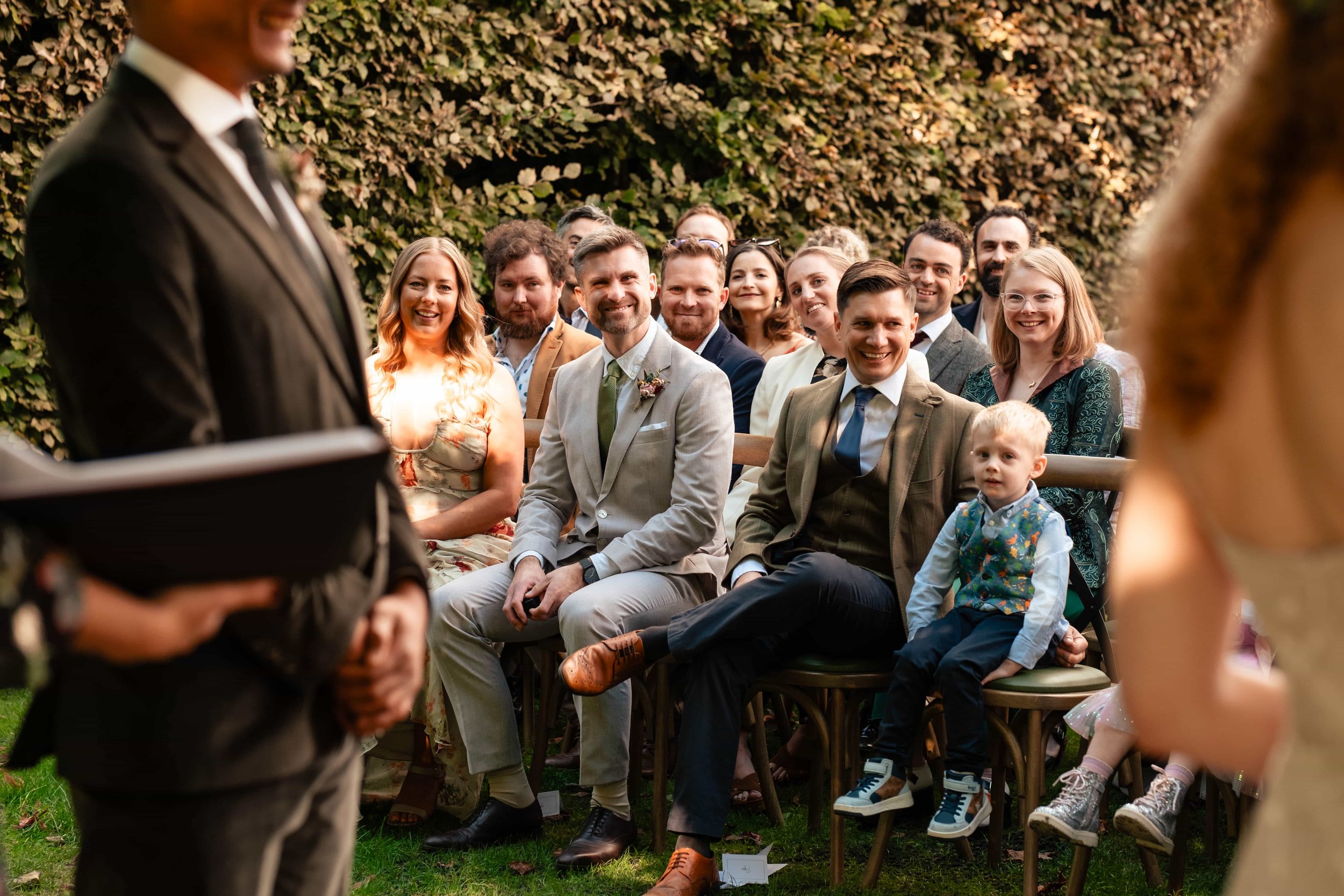 People sitting on chairs outdoors during a wedding ceremony, smiling and watching a person in a suit speaking. Green hedge in the background.