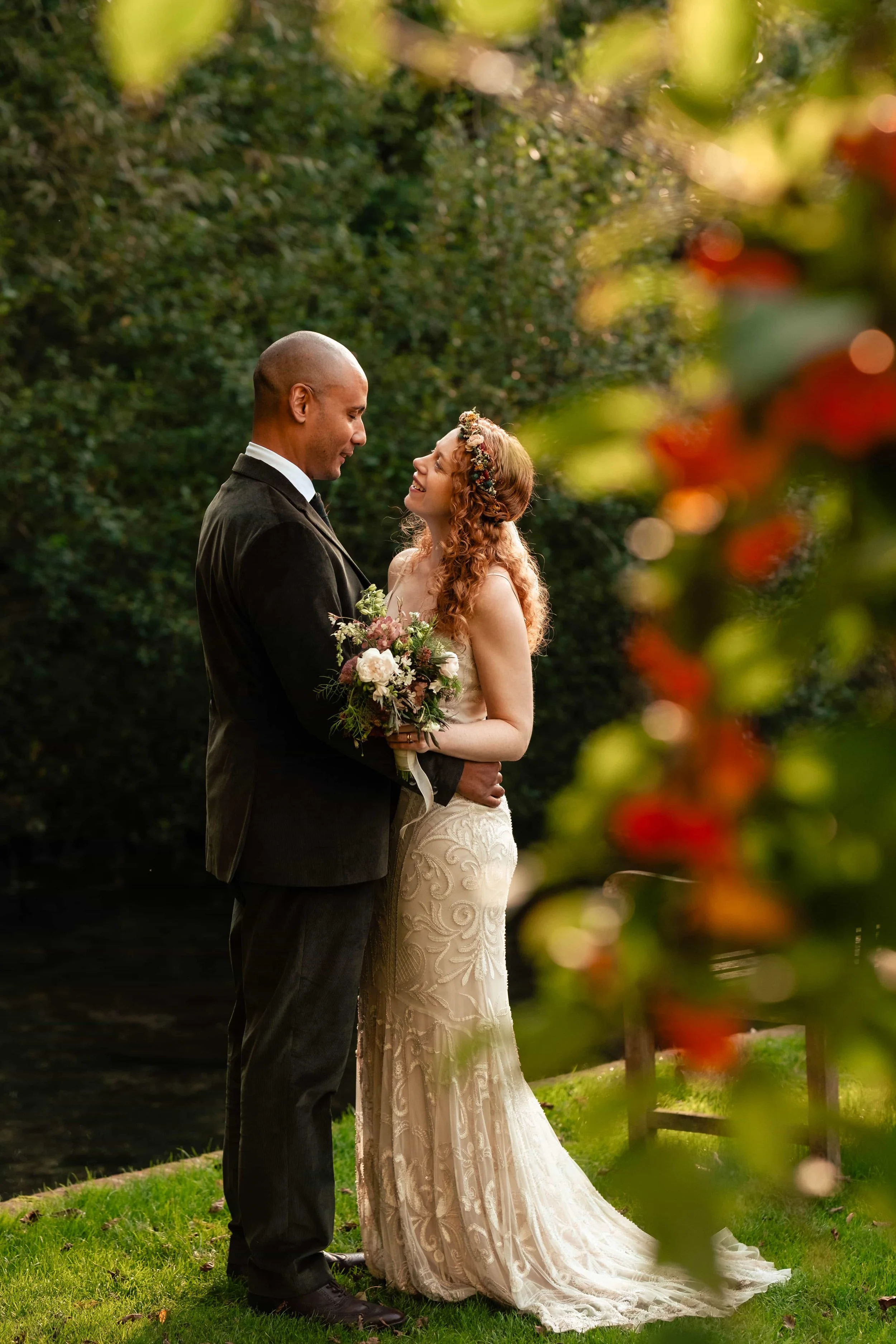 A newlywed couple stands face-to-face outdoors, smiling at each other. The man is dressed in a dark suit and white shirt, holding a bouquet, while the woman wears a lace wedding gown and a floral headband. The background features greenery, and blurre