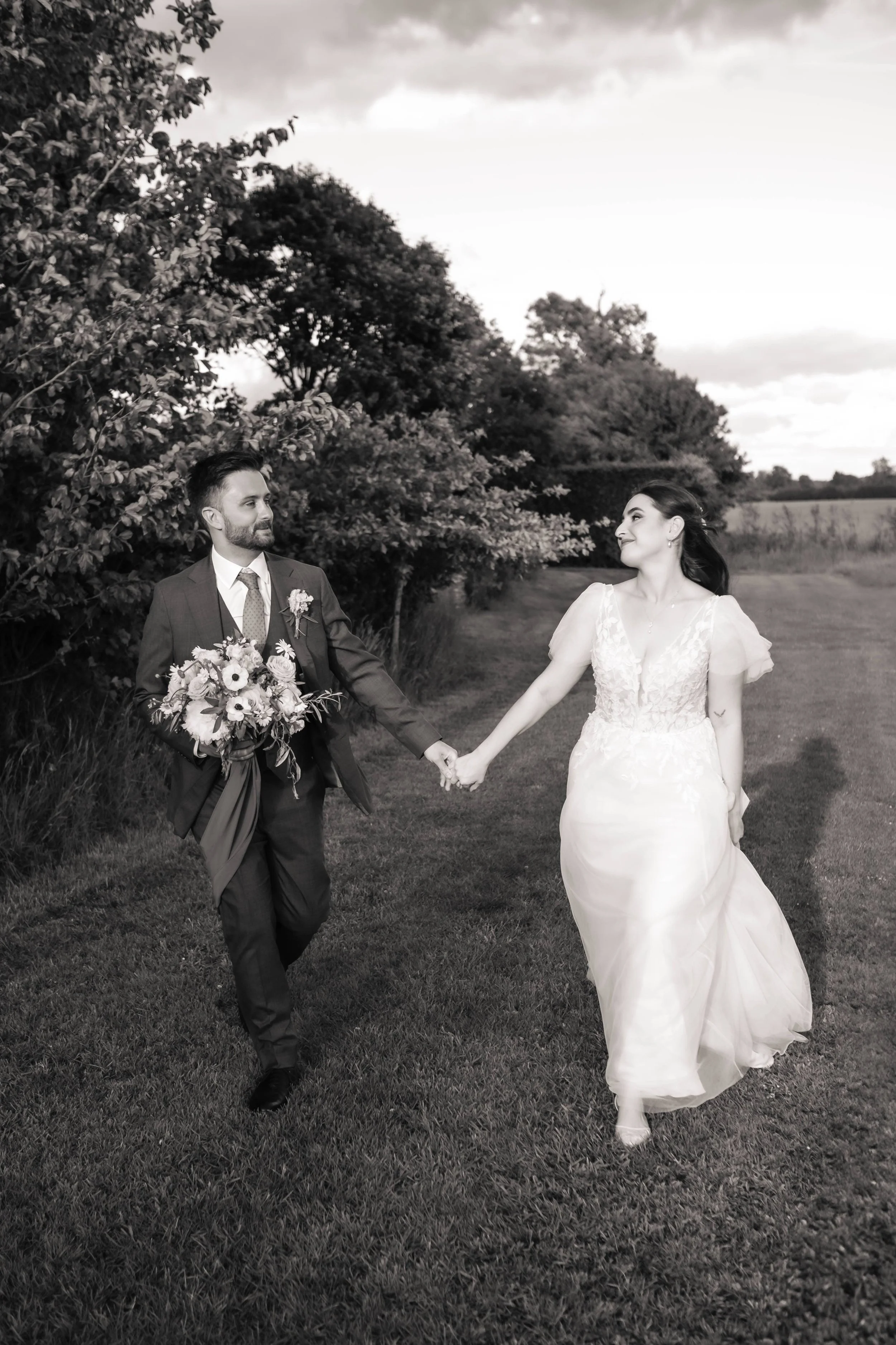 Black and white photo of a bride and groom holding hands and walking on a grassy field outdoors, with trees and a cloudy sky in the background.