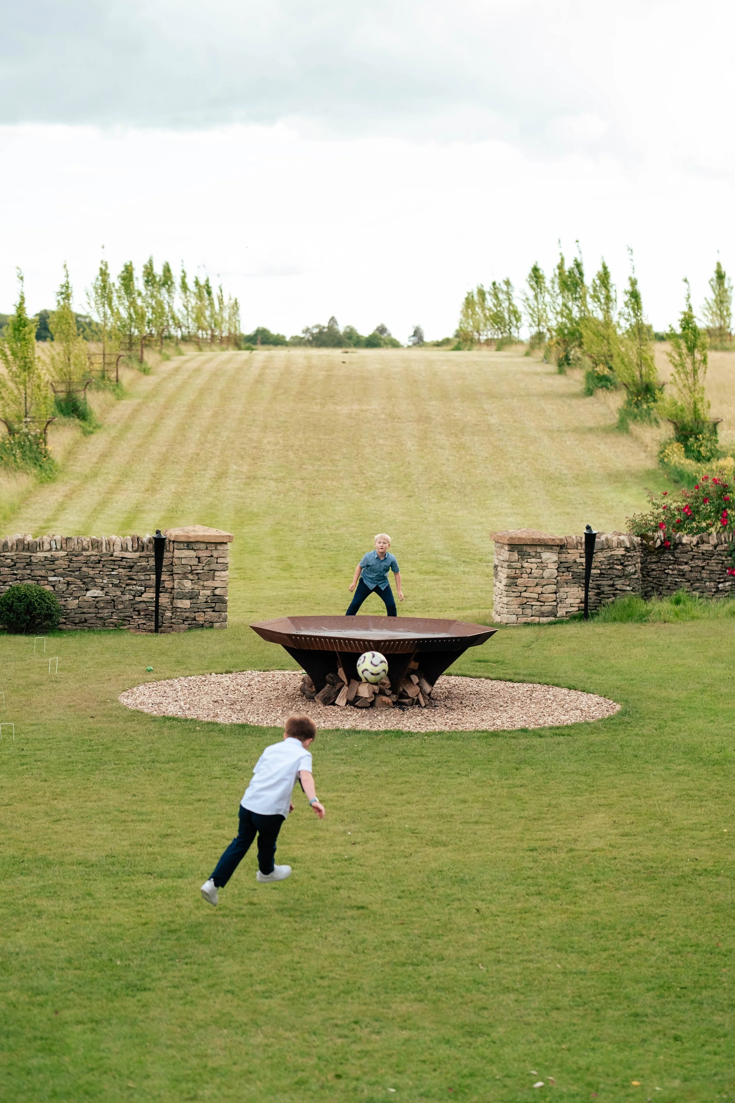 Two boys playing soccer on back lawn of Cripps Barn near a fire pit with a decorative soccer ball inside it, against a backdrop of a hill with trees and a cloudy sky.