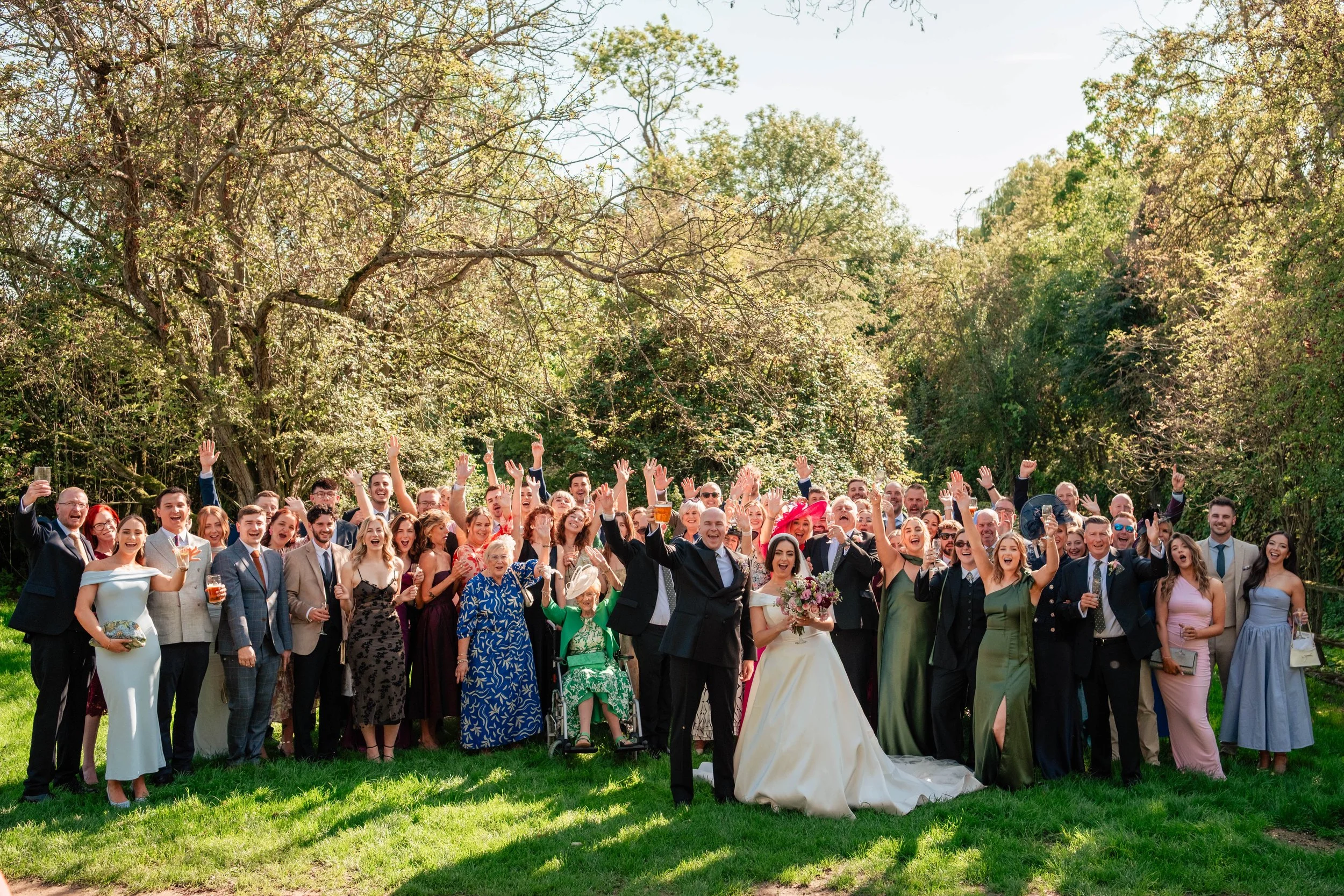 A large group of people gathered outdoors on a grassy area, celebrating a wedding. The group includes men, women, and elderly individuals, all dressed in formal attire, with some holding drinks. In the center, a woman in a white wedding dress holding