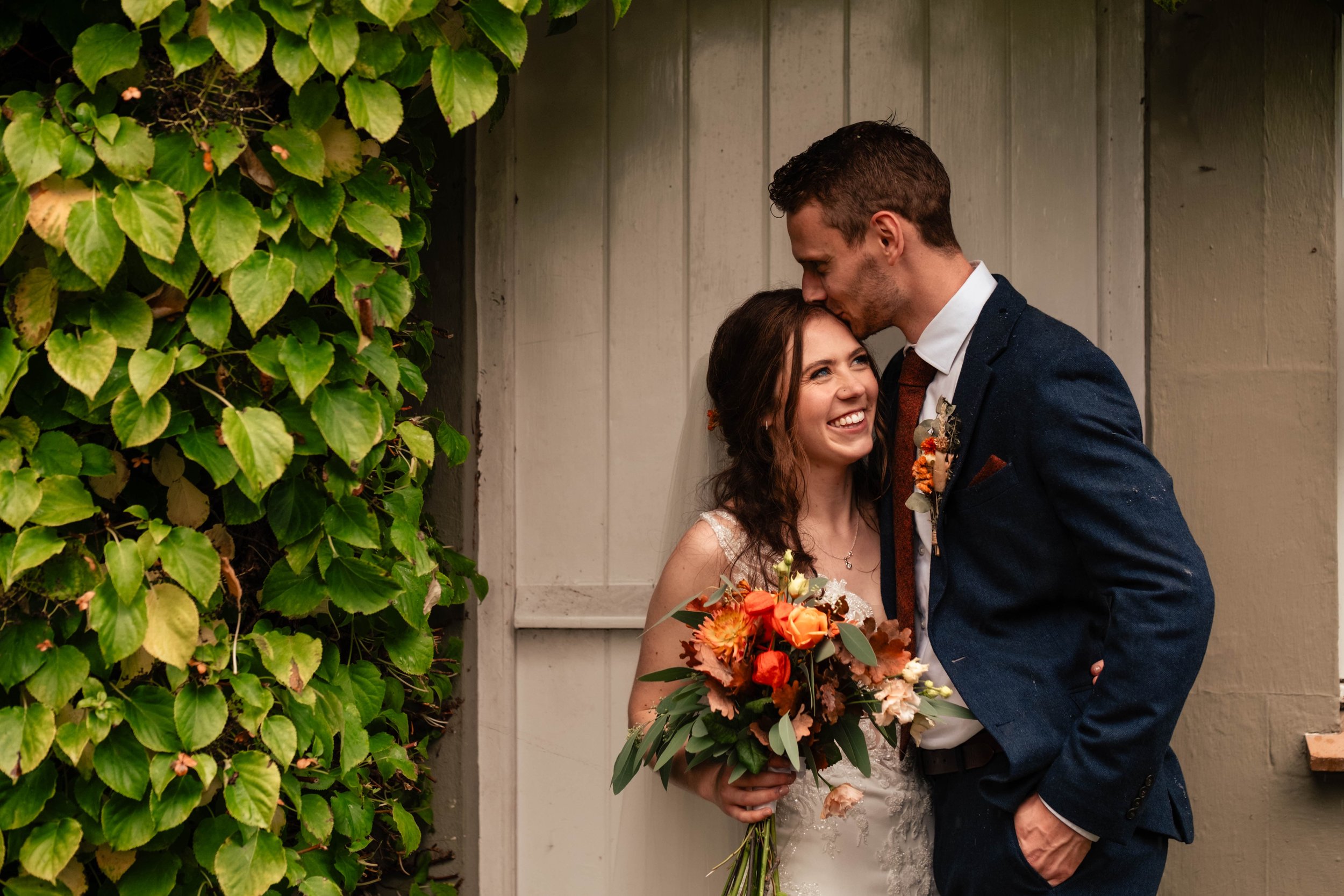 A smiling bride holding a bouquet of flowers and a groom kissing her on the forehead, standing beside a beige wooden wall and green leaves.