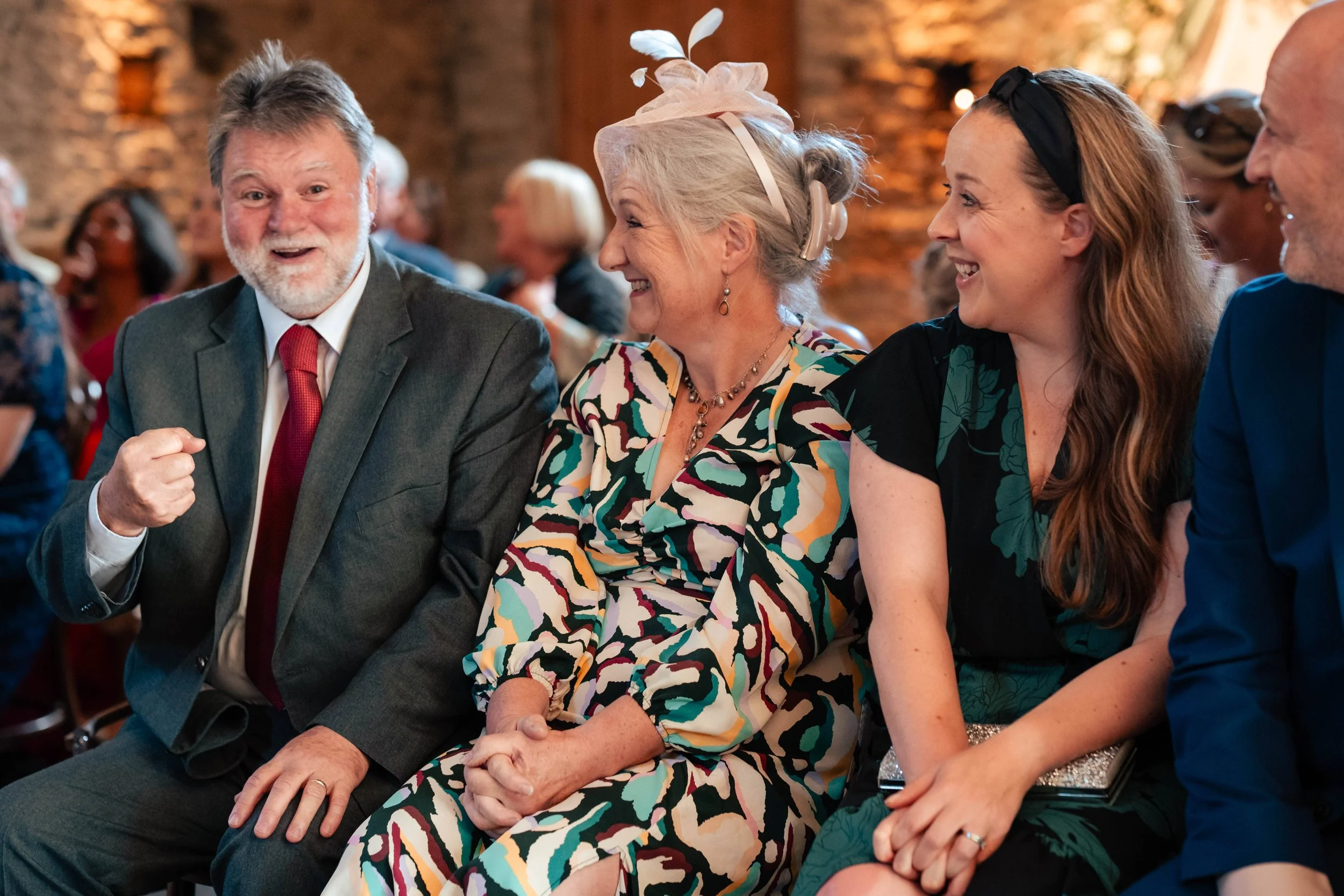 A group of four people sitting together at a social event, smiling and engaging in conversation. The man on the left is wearing a gray suit with a red tie, the older woman next to him is dressed in a multicolored patterned dress with a pink hat and b