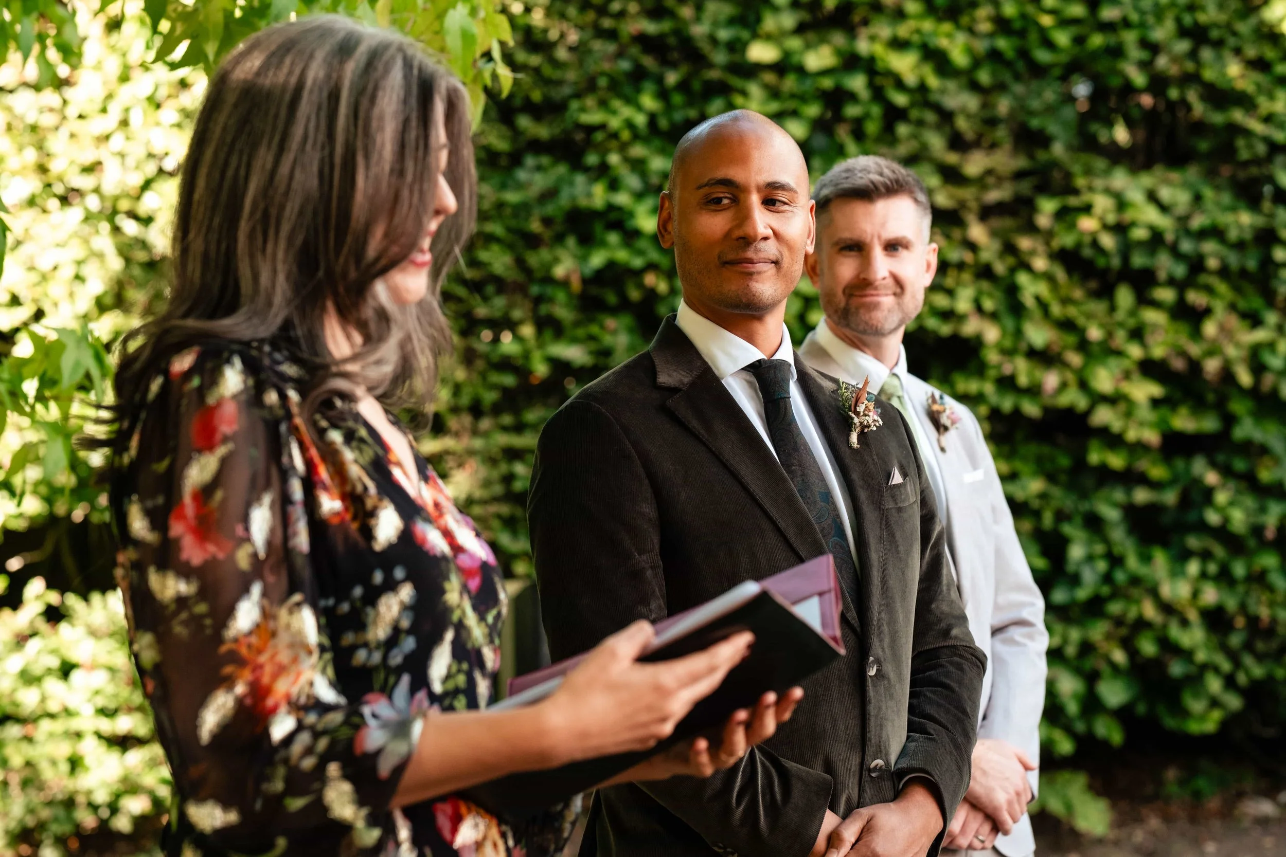 A wedding ceremony outdoors with three people standing in front of a green hedge. The officiant is on the left, holding a book and facing the groom, who is in the center, dressed in a dark suit. The best man is on the right, dressed in a light suit. 