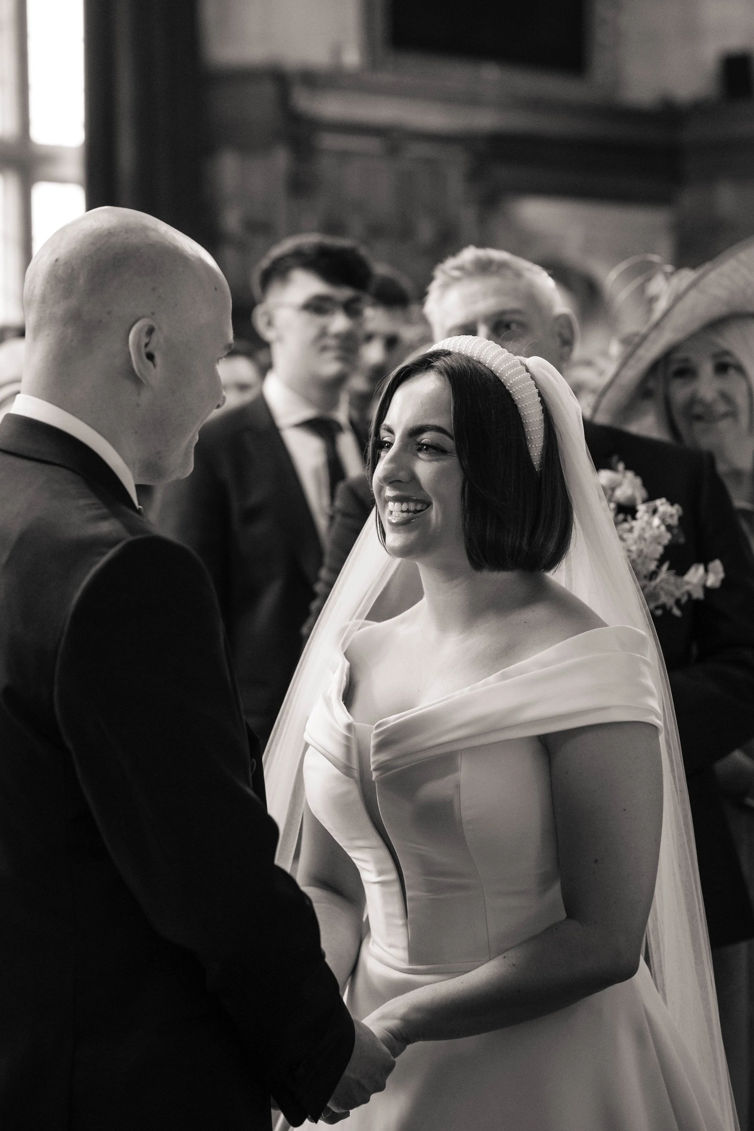 A black and white photo of a bride and groom holding hands during their wedding ceremony, smiling, with guests watching in the background.