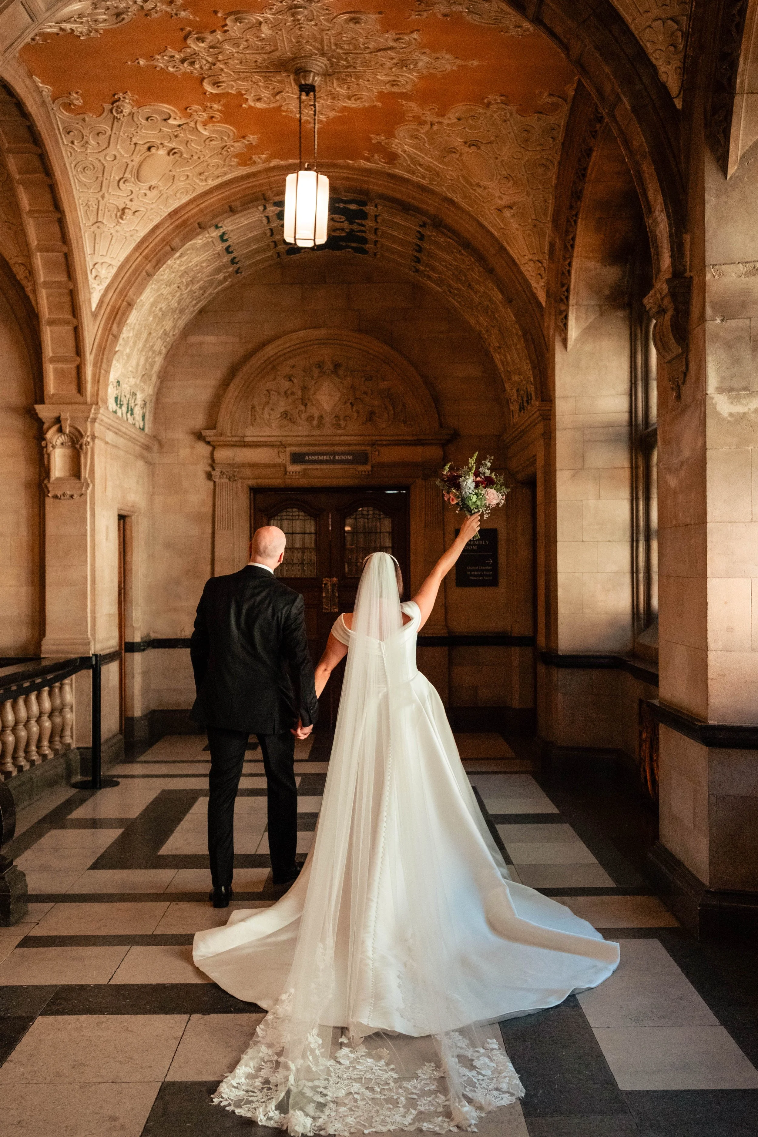 A bride in a white wedding gown and veil holding a bouquet of flowers, with her arm raised, walking hand-in-hand with a groom in a black suit down a grand hallway with ornate ceiling and arched doorway.