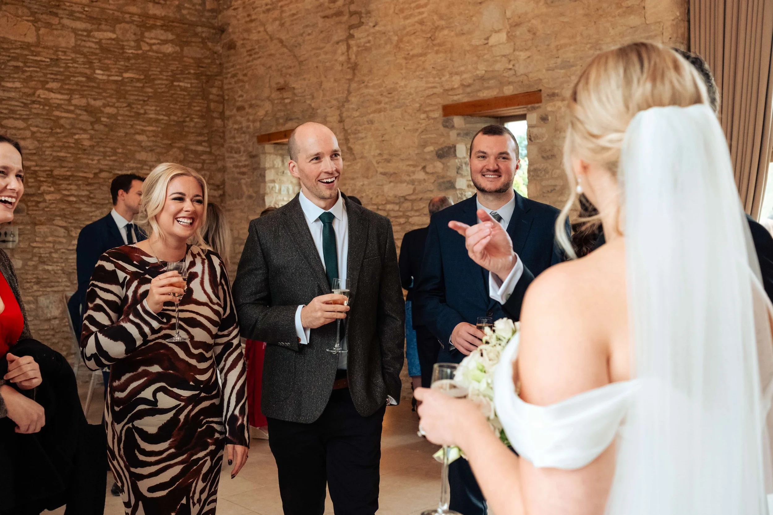People at a wedding reception, standing in a room with brick walls, celebrating and talking, with some holding glasses of champagne.