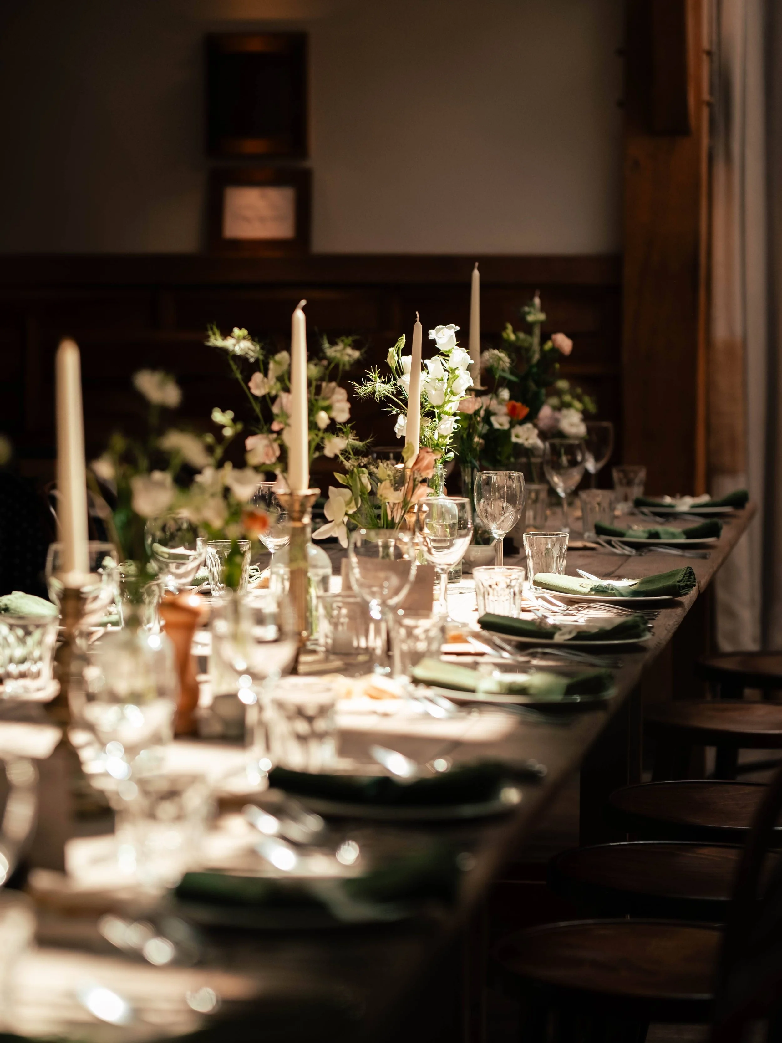 Elegant dining table set with white flowers, tall candles, green napkins, and glassware in dim lighting, likely prepared for a special occasion.