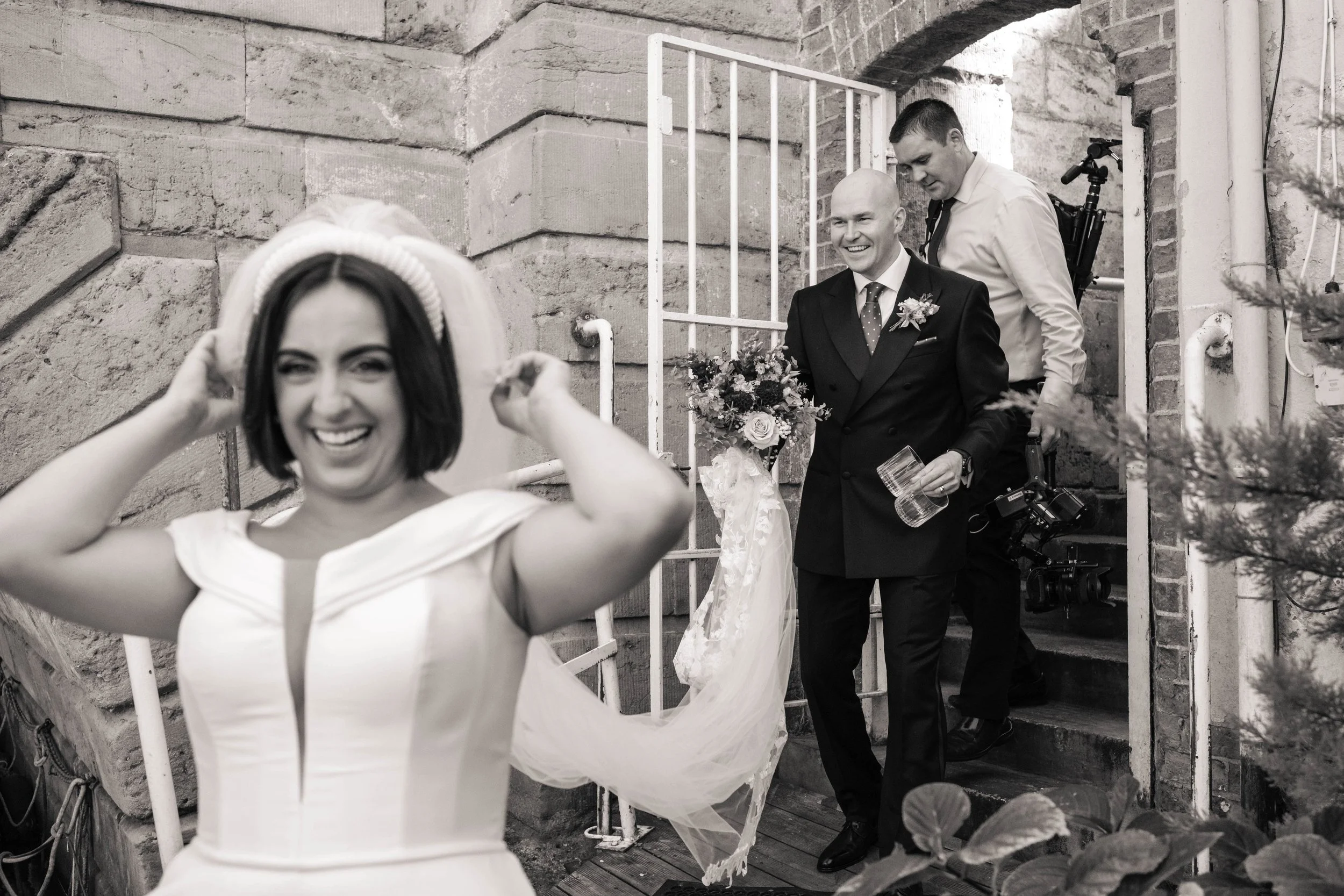 A bride with short hair and a dress adjusting her veil while smiling, with two men descending stairs behind her, one holding a bouquet of flowers and a glass, and the other with a camera on his back, in a brick outdoor setting.