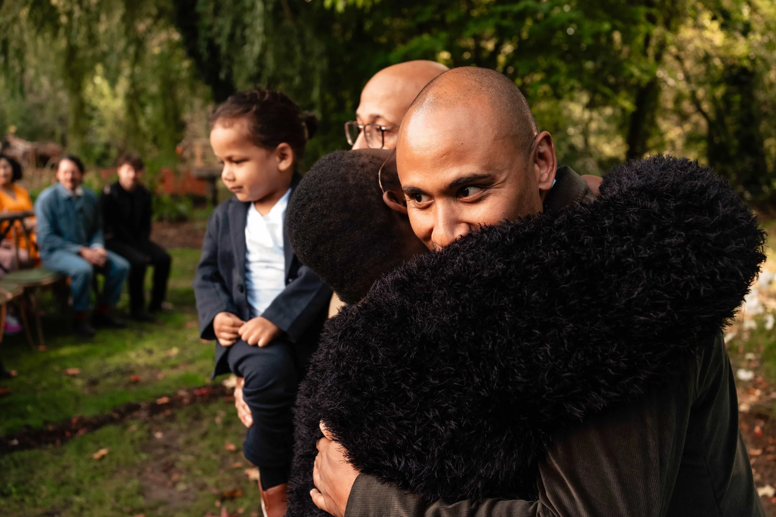 A man embracing a woman outdoors with children and other adults in the background.