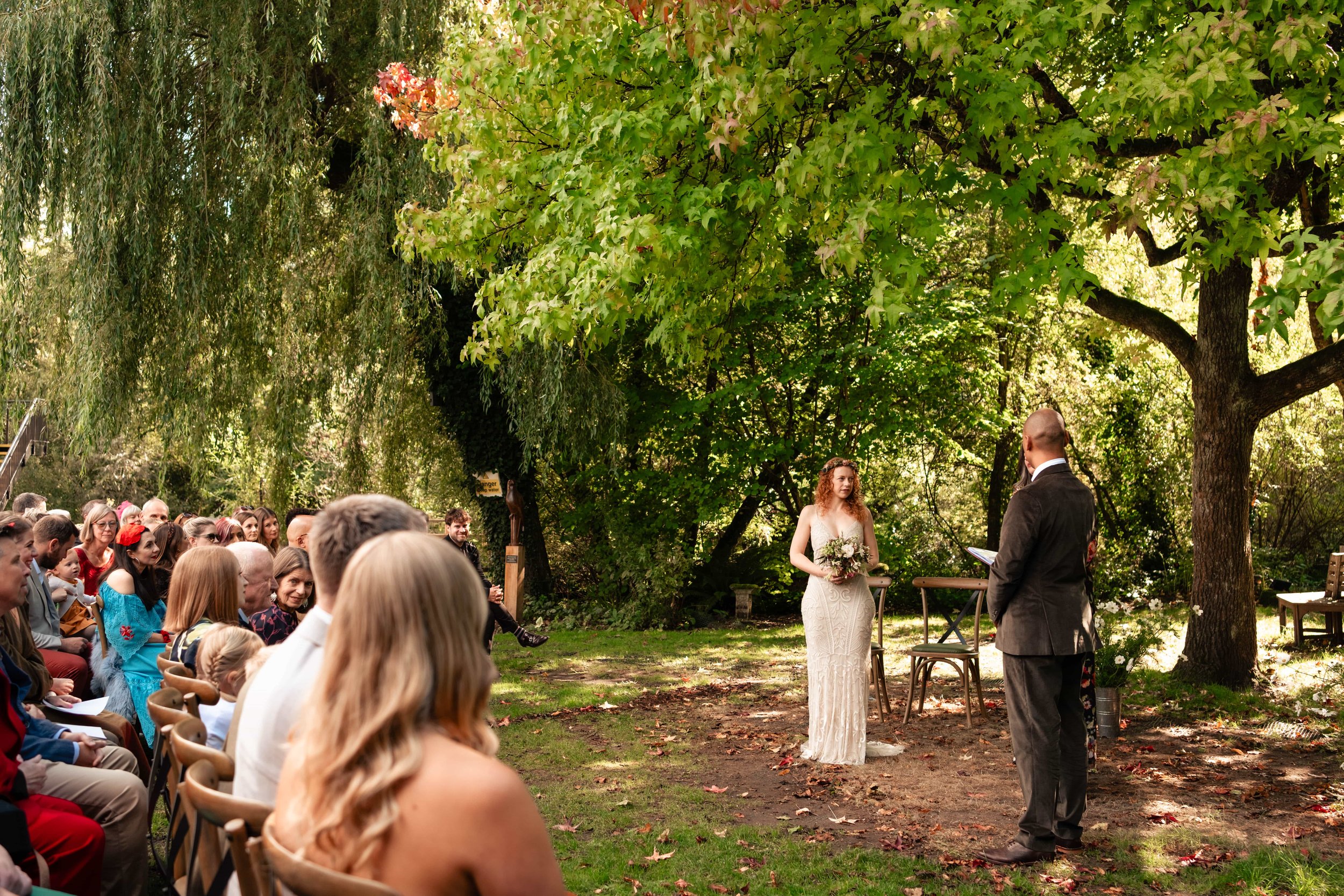 A wedding ceremony taking place outdoors in a lush green garden, with a bride holding a bouquet and a groom facing her, surrounded by seated guests.