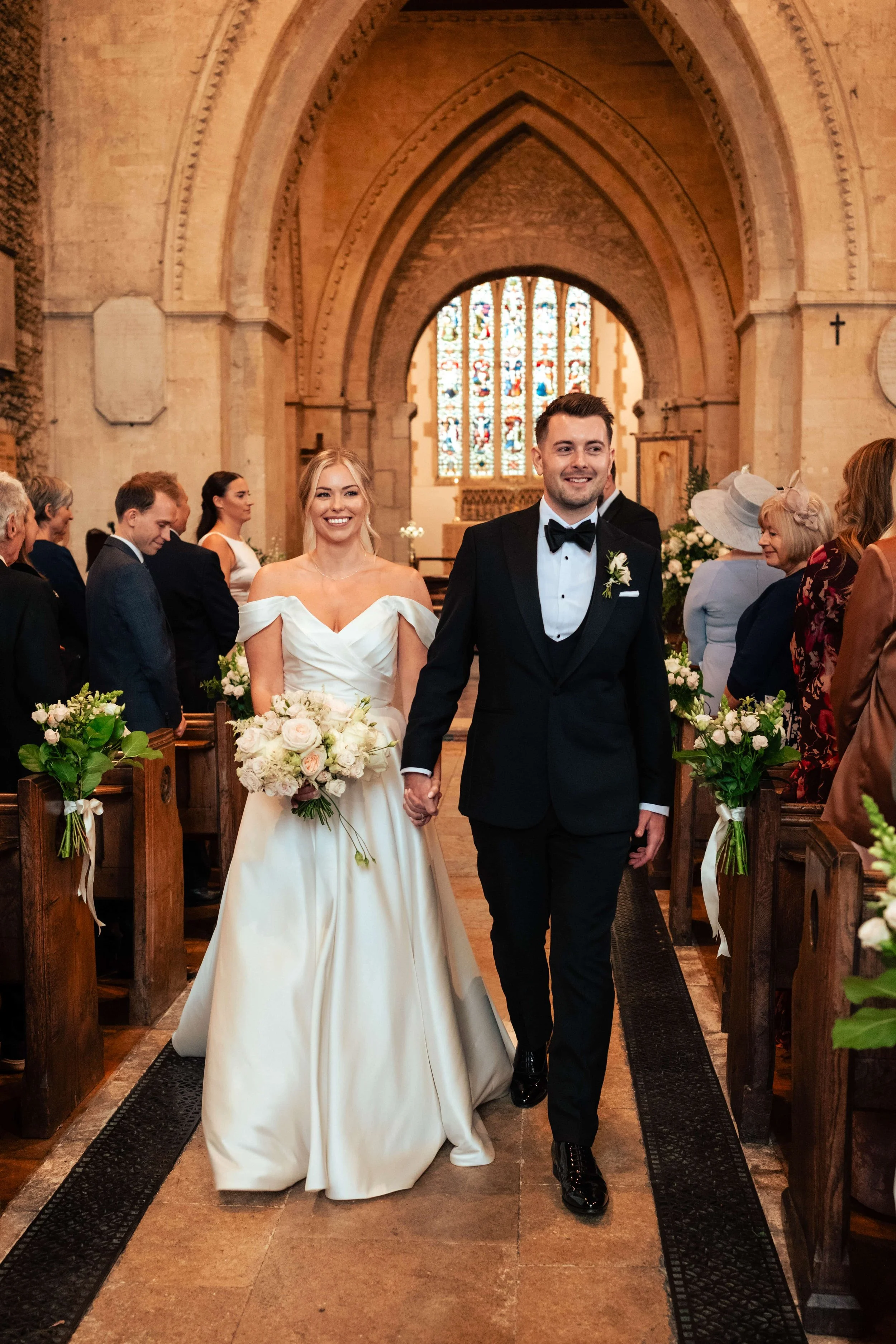A bride and groom walking hand in hand down the aisle in a church, surrounded by wedding guests, with stained glass windows in the background.