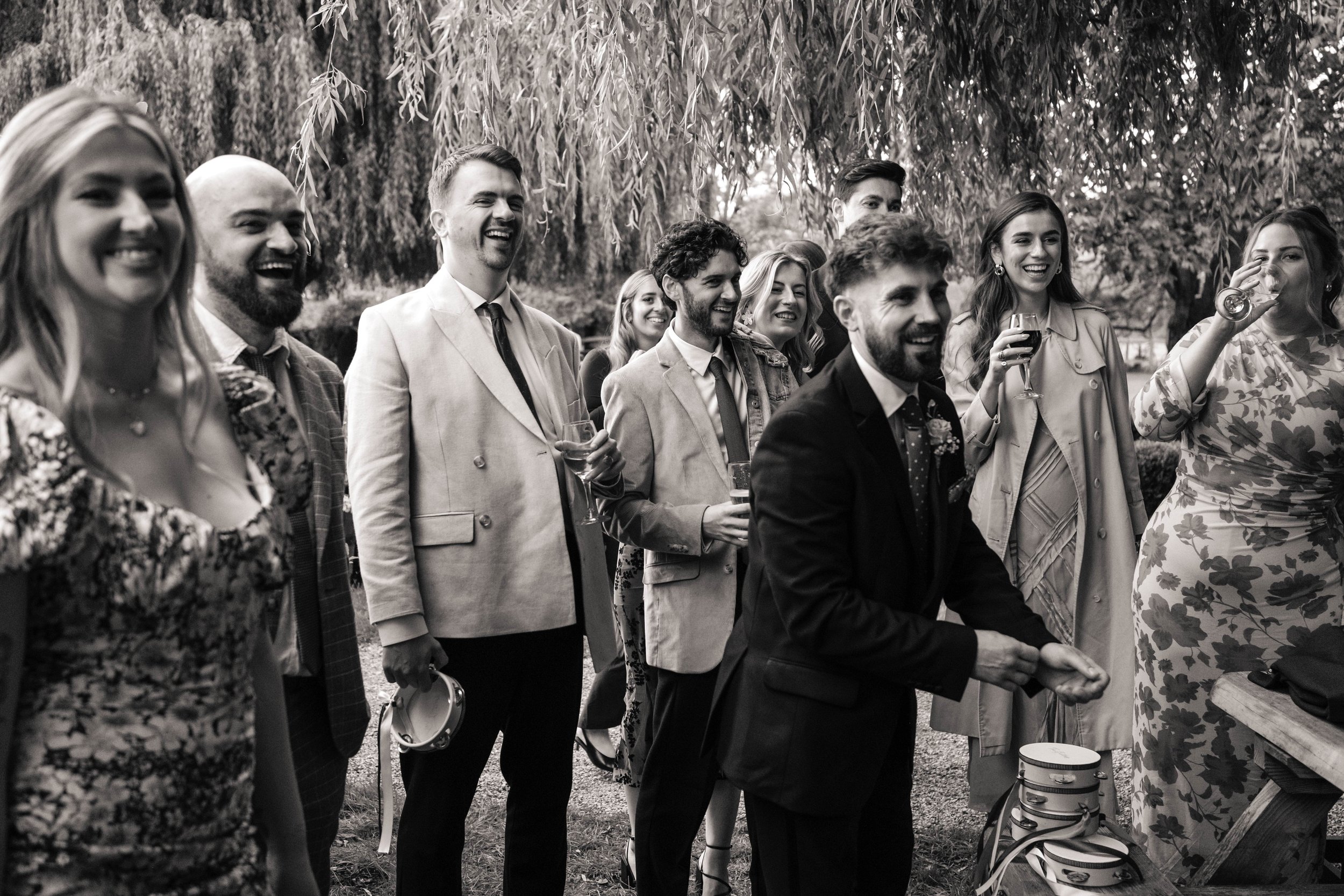 Group of people enjoying an outdoor party, some holding drinks, under trees, black-and-white photo.