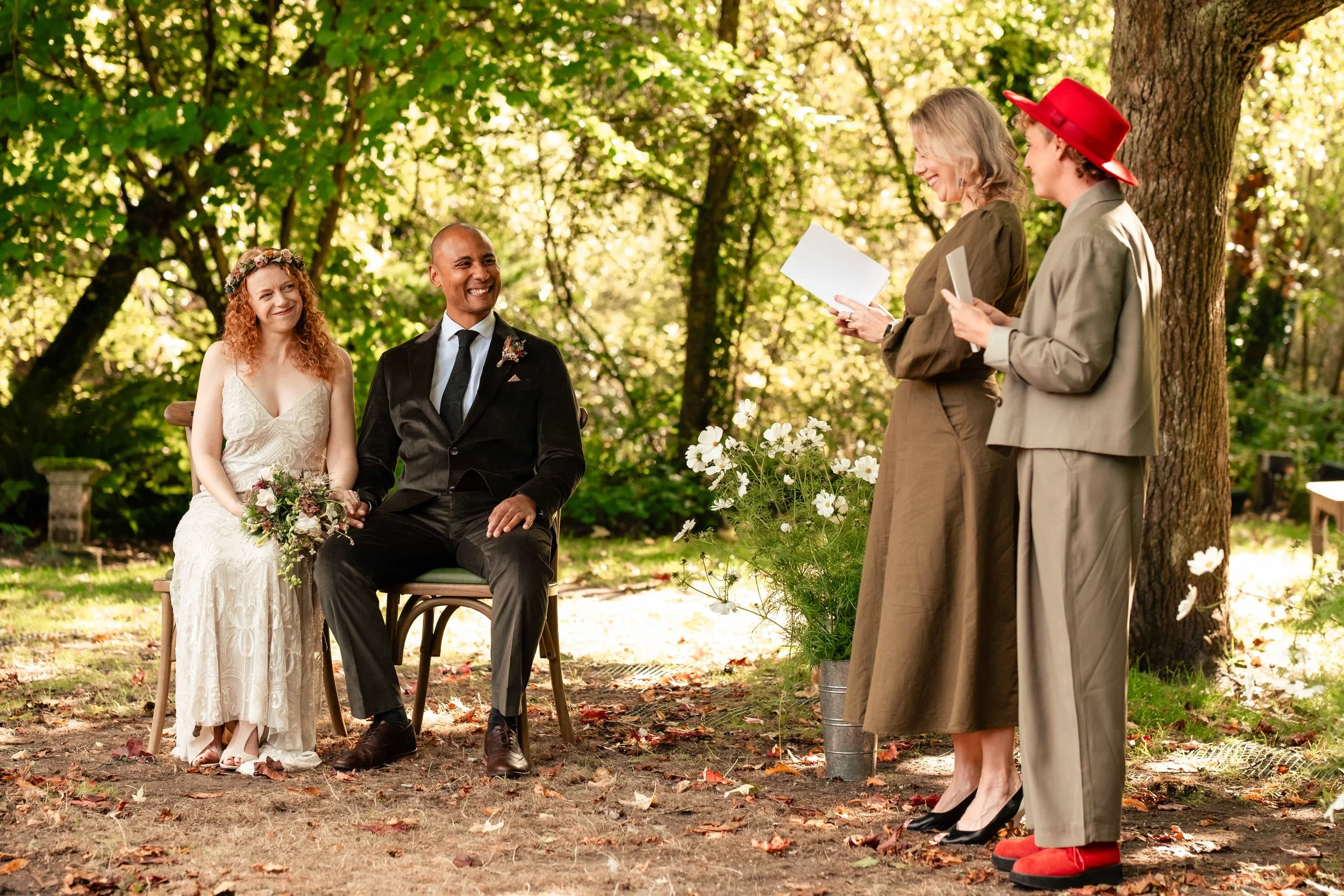 A wedding ceremony outdoors with a bride and groom sitting on a bench, holding hands and smiling. The bride is wearing a white lace dress and holding a bouquet, and the groom is in a black suit. Two women are standing in front of them, reading from p