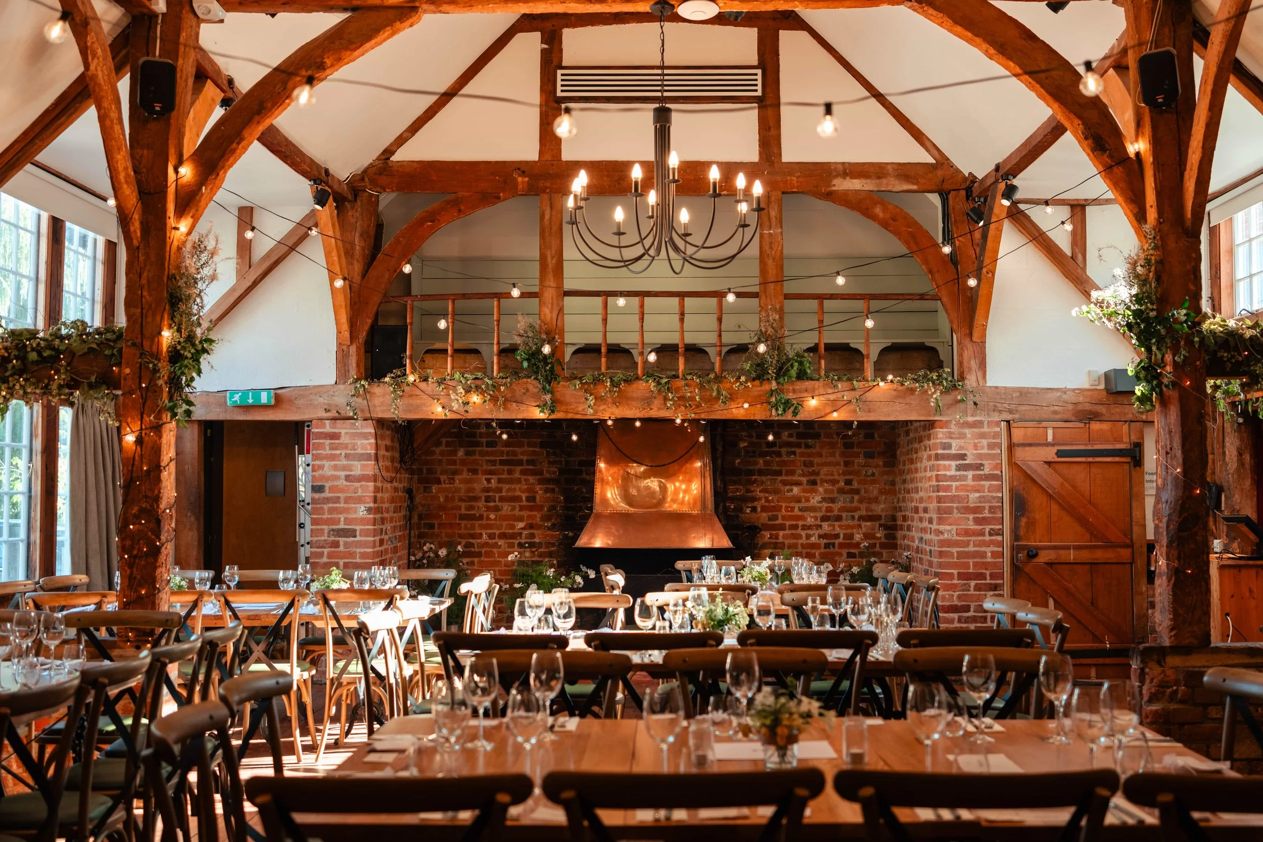 Beautifully decorated rustic wedding reception hall with wooden beams, string lights, and a chandelier, featuring long tables set with glassware and floral centerpieces, brick wall, and a balcony area.