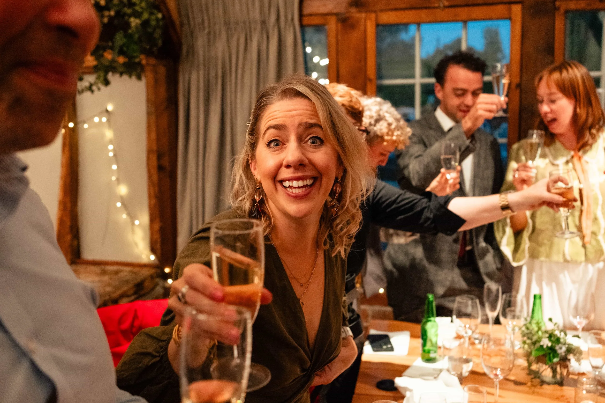 People celebrating at a dinner party, toasting with drinks, including a smiling blonde woman in the foreground.