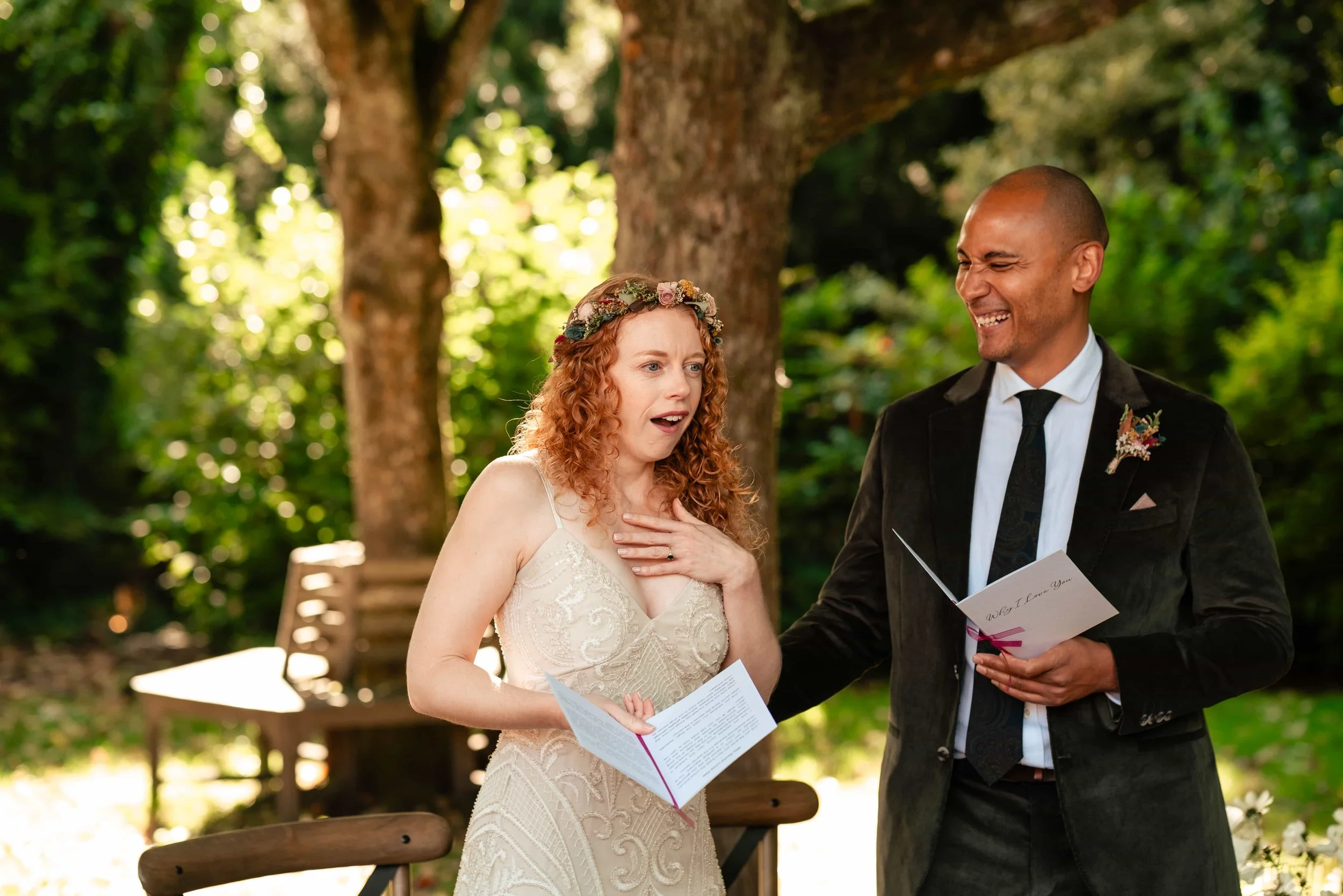 A wedding ceremony outdoors with a woman in a beige lace dress and a flower crown looking surprised, and a man in a black suit and tie smiling, holding a card in a garden with trees and benches in the background.