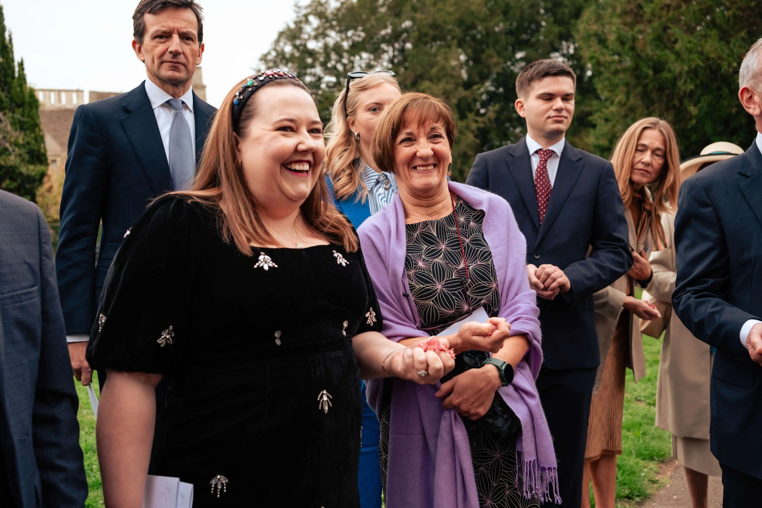 A group of six people standing outdoors, smiling, with trees in the background. The group includes three women and three men dressed in formal attire.