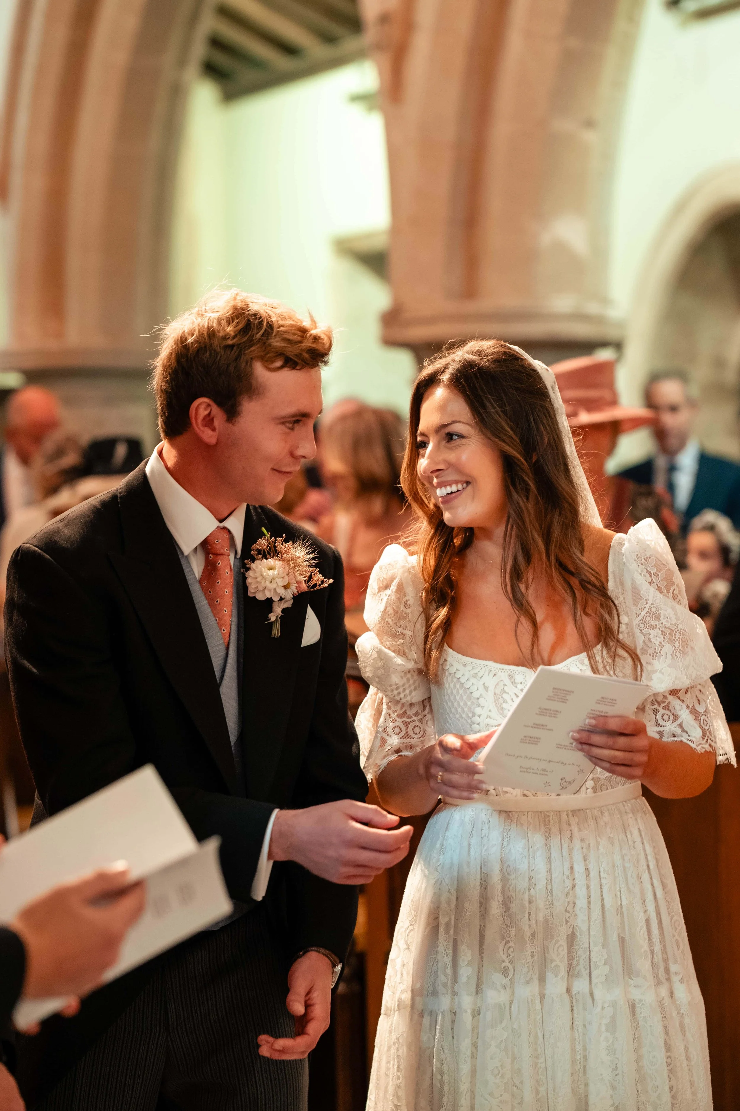 A bride and groom sharing a moment during their wedding ceremony inside a church, with the bride holding a program and smiling at the groom.