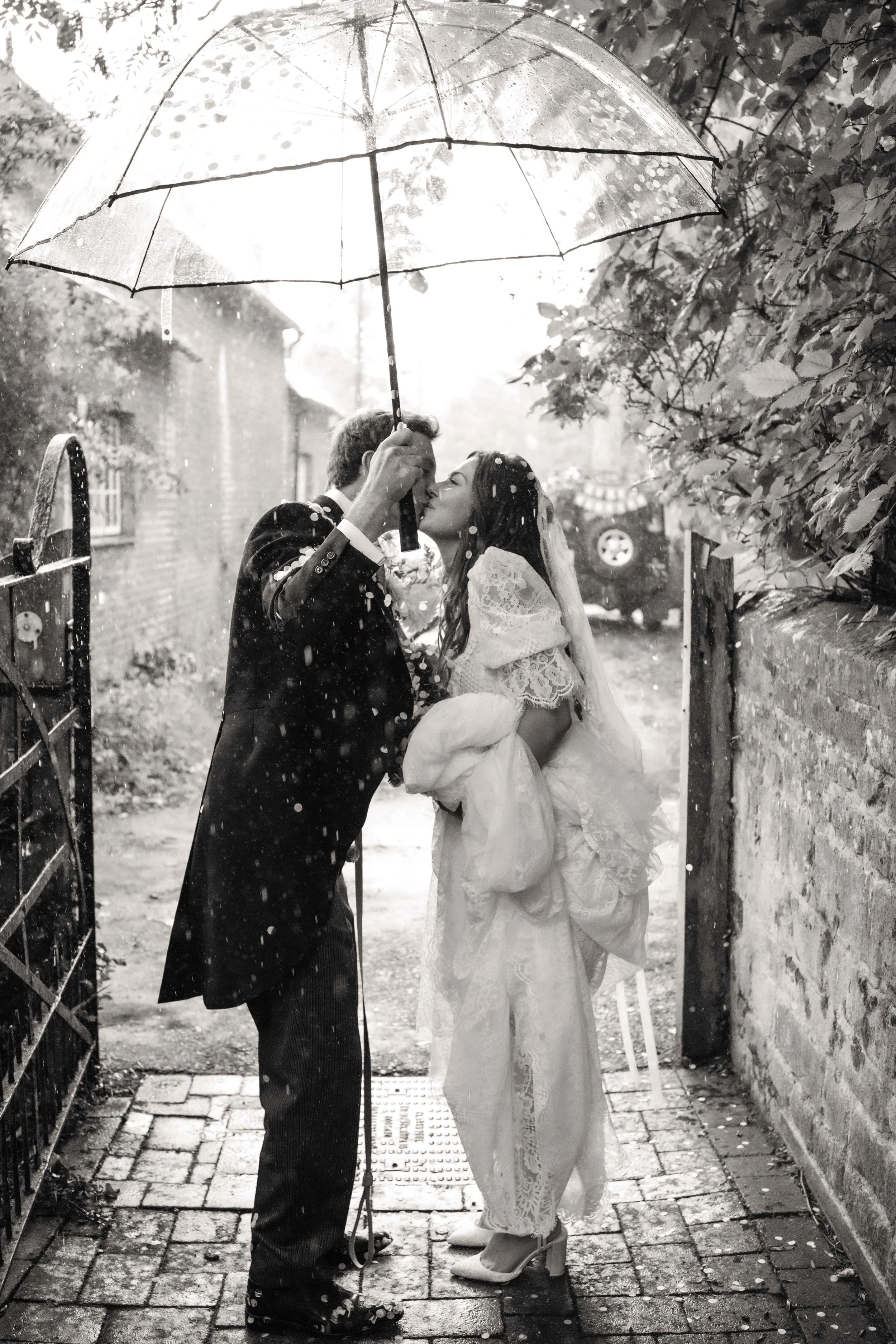 A couple shares a romantic moment under an umbrella in the rain, with the man kissing the woman's forehead.