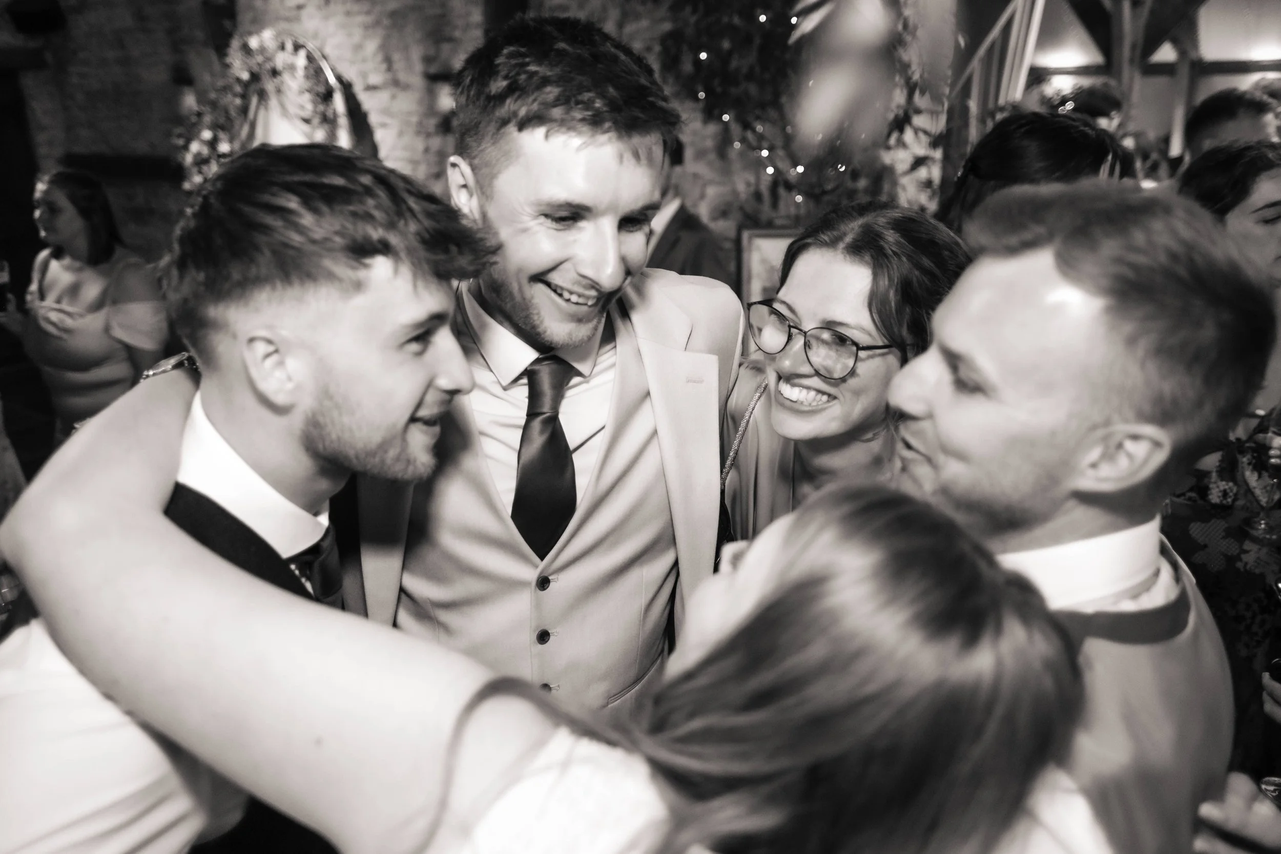 Group of five people, four men and a woman, smiling and hugging at a celebration or social gathering in a dimly lit venue.