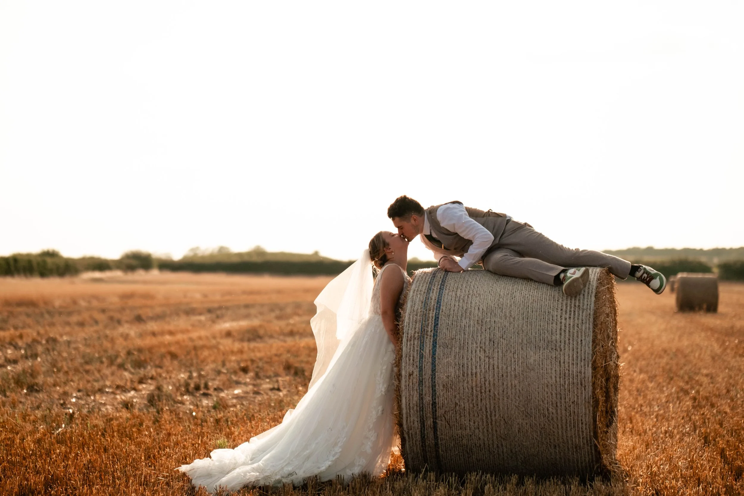 A bride in a white wedding dress and veil standing on the ground, and a groom in a gray suit lying on top of a large hay bale in a field during sunset, touching noses.