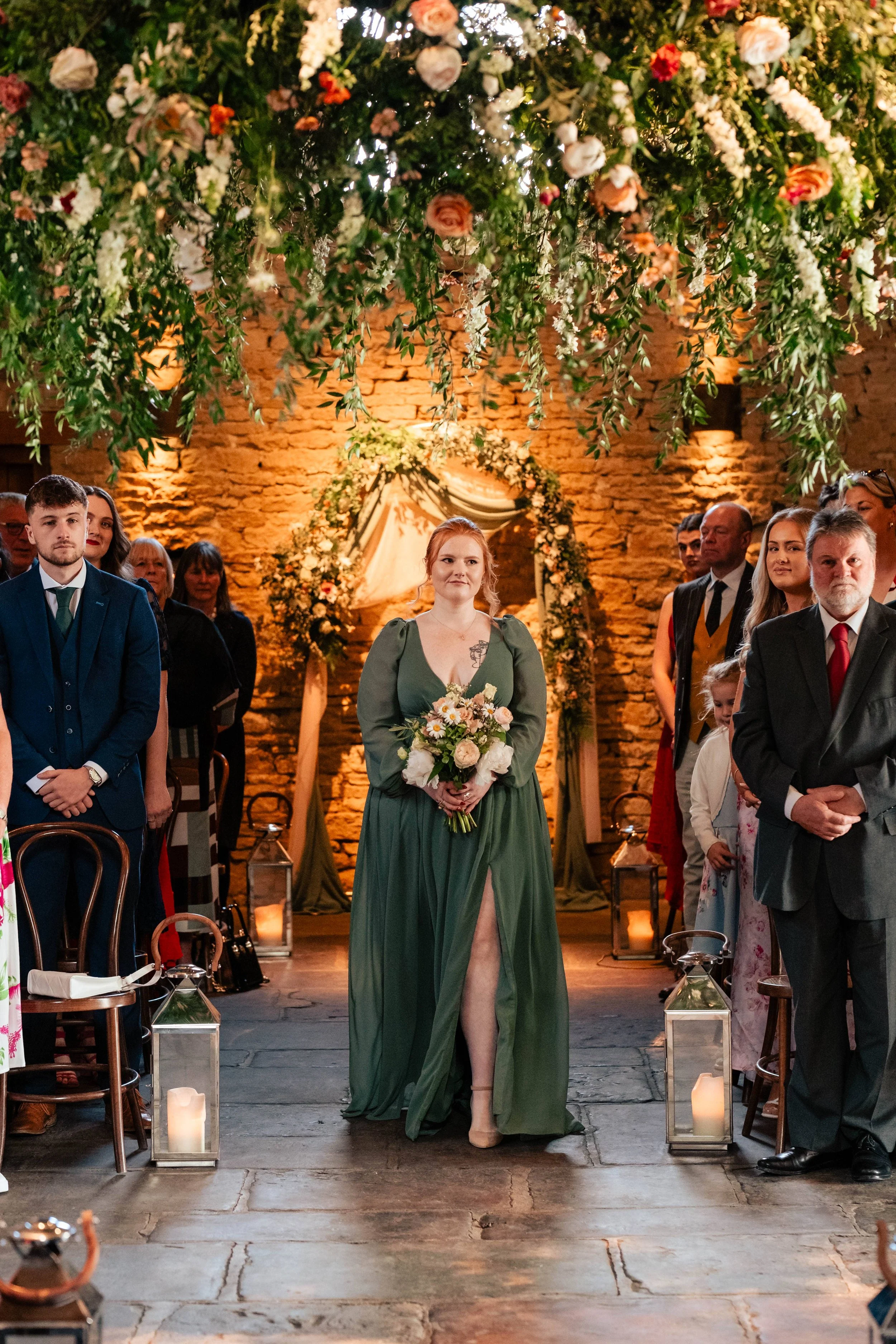 A woman in a green dress holding a bouquet of flowers, standing at a wedding ceremony in a rustic indoor venue with exposed brick walls and floral decorations.