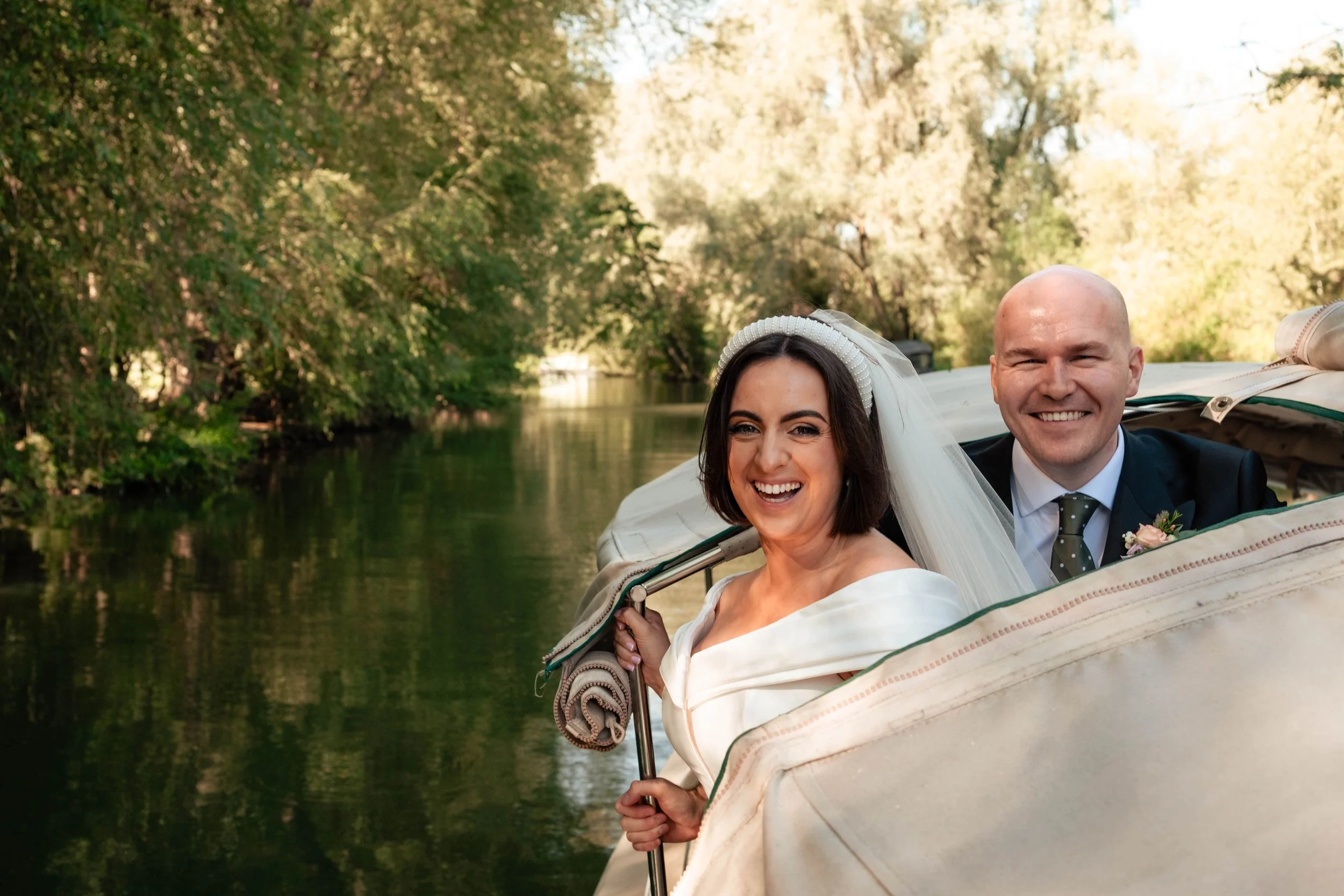A bride and groom in wedding attire sailing on a boat through a lush, green river surrounded by trees, smiling joyfully.