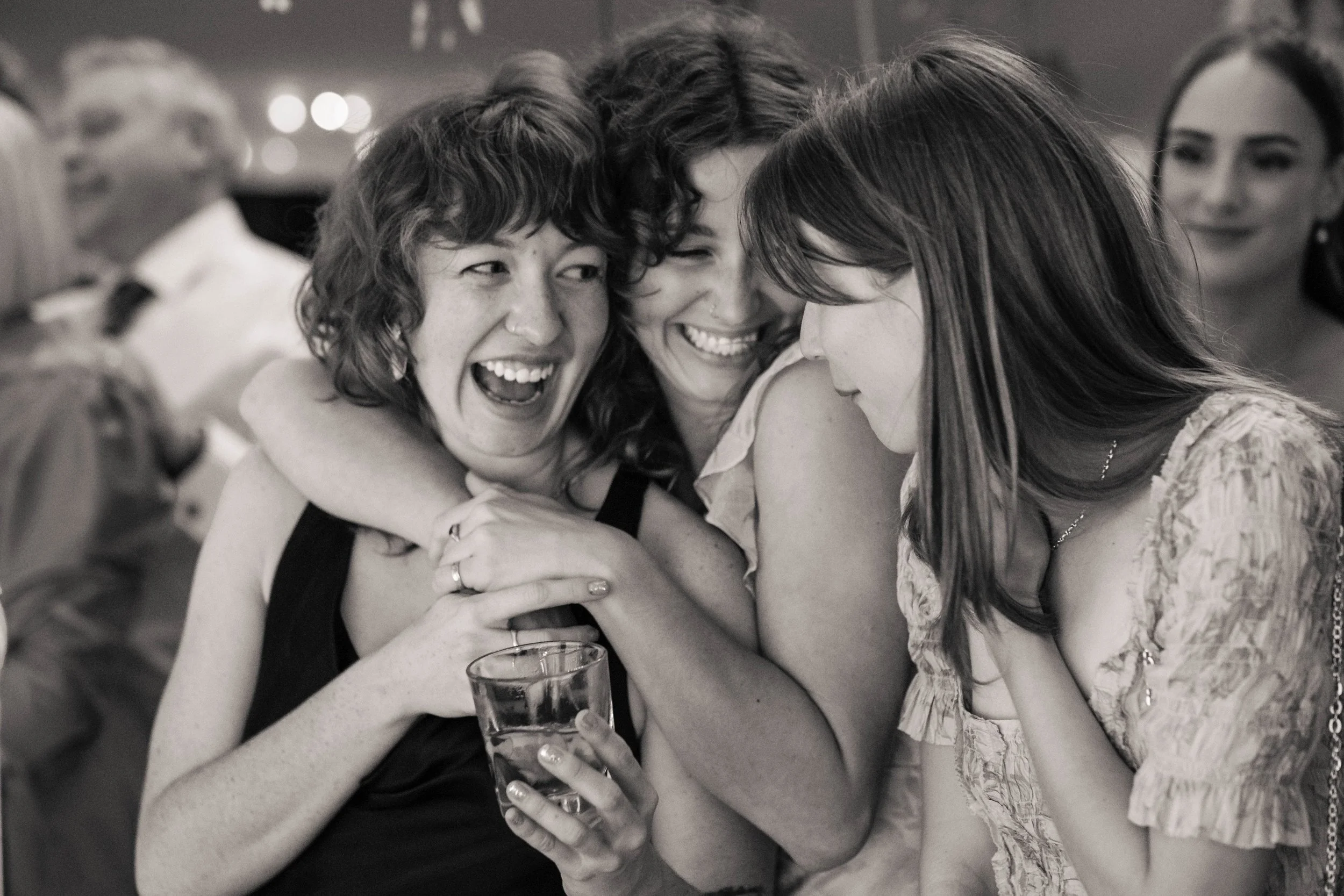 Four women huddled together smiling and laughing at a social gathering, one holding a drink, in black and white.