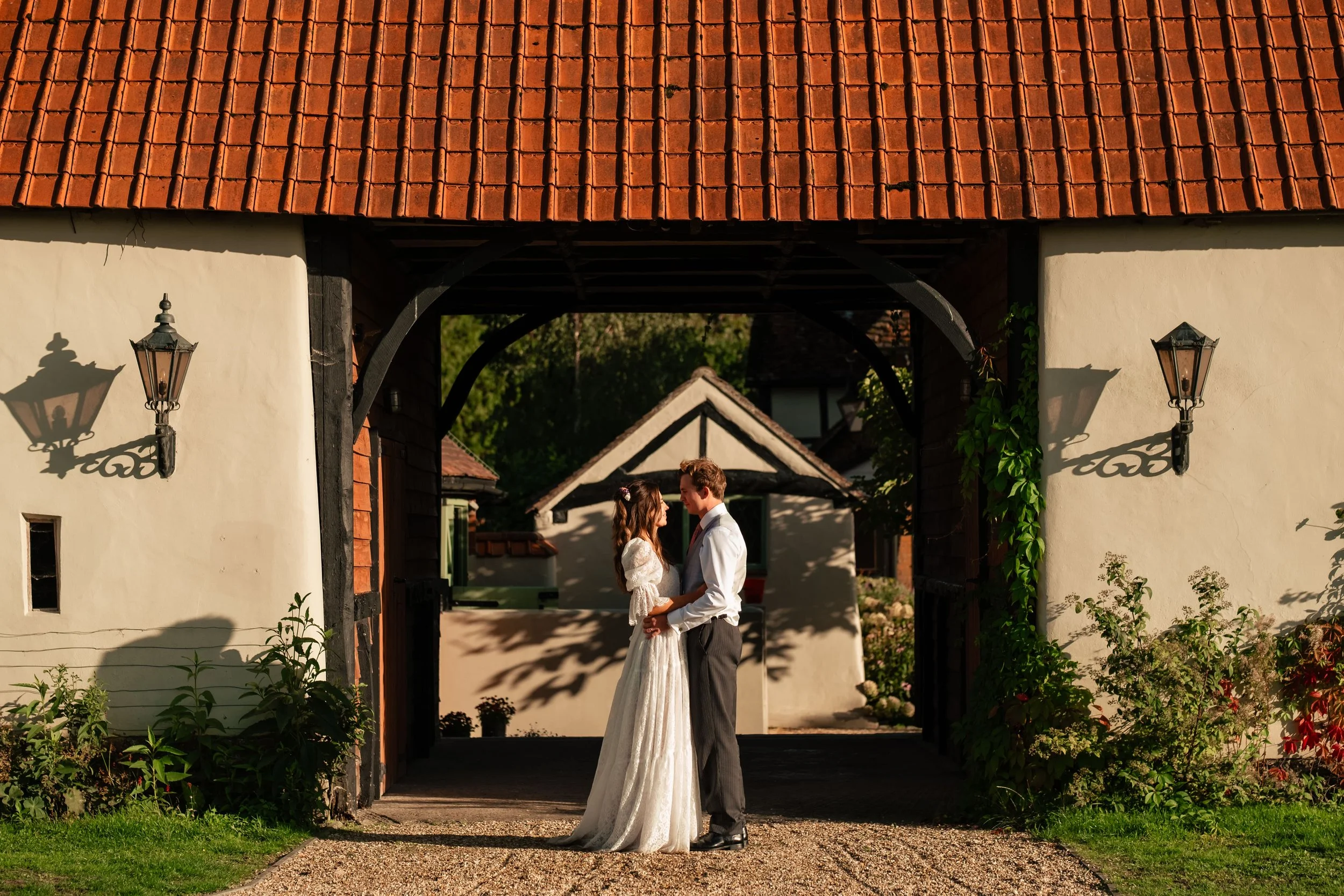 A newlywed couple stands close together under an archway of a rustic cottage, facing each other, with greenery and small buildings in the background.