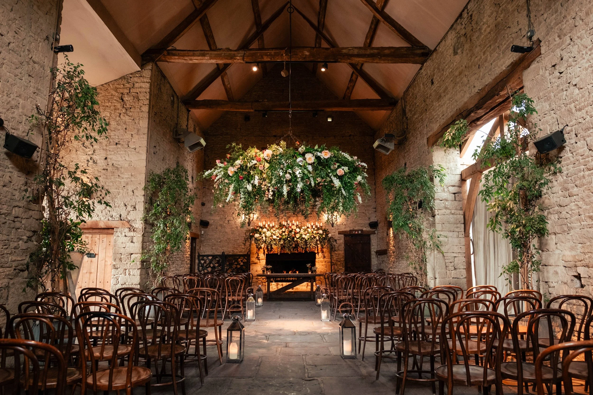 Rustic wedding ceremony setup in a barn with wooden chairs, floral arrangements, candles in lanterns along the aisle, and a large floral chandelier hanging from the ceiling.