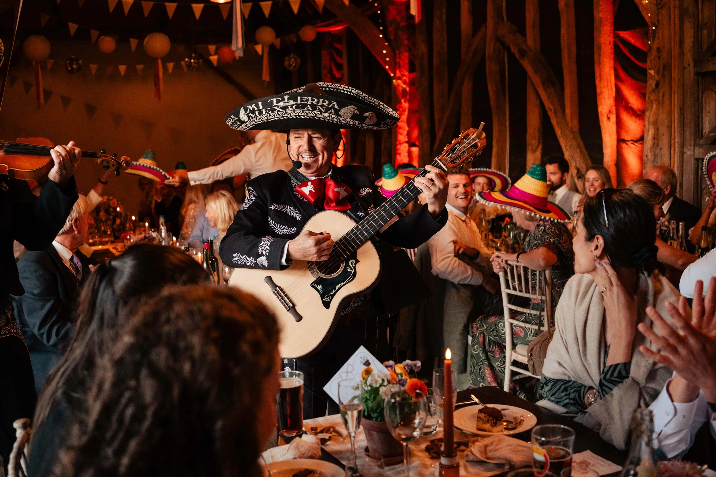 A wedding celebration with a man wearing a traditional sombrero and playing a guitar, surrounded by people wearing colorful sombreros, in a festive decorated barn-like setting.