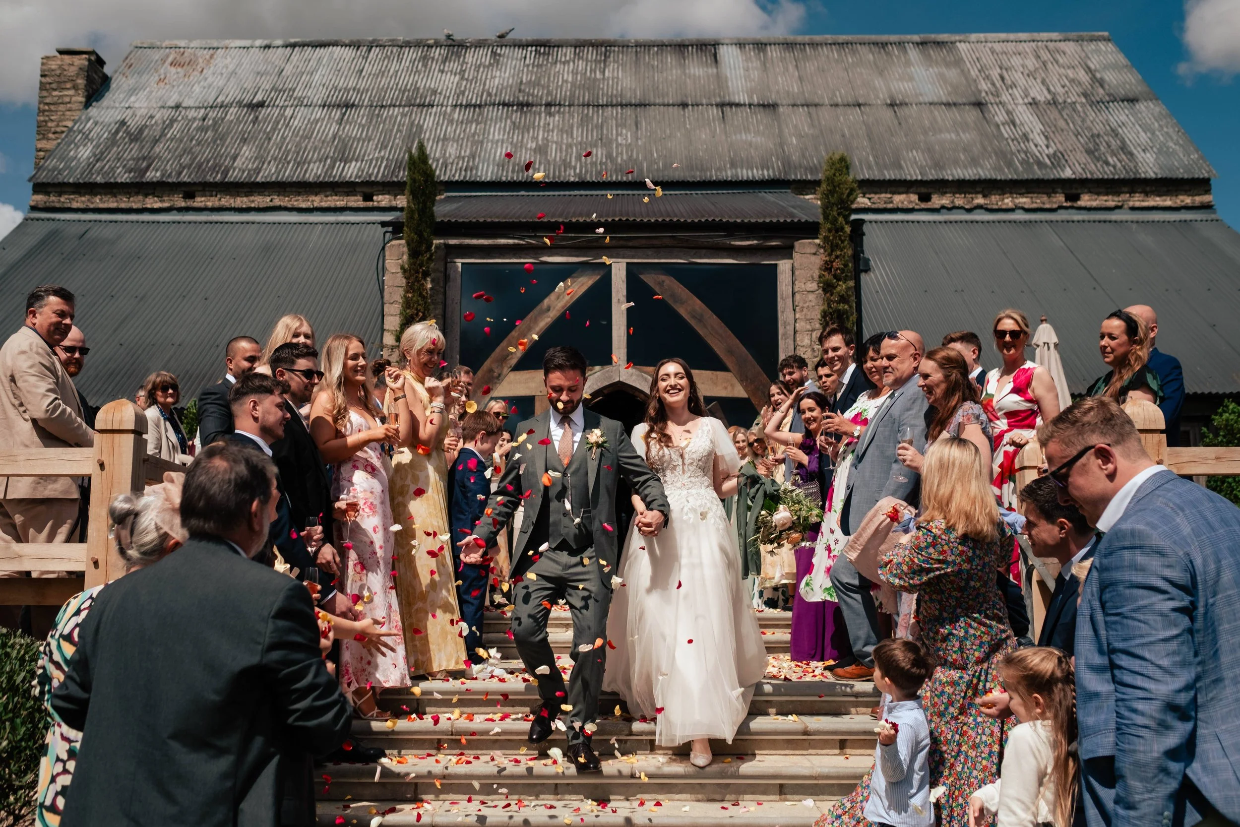 A newlywed couple walking down the steps of a rustic venue with family and friends celebrating with flower petals thrown in the air.